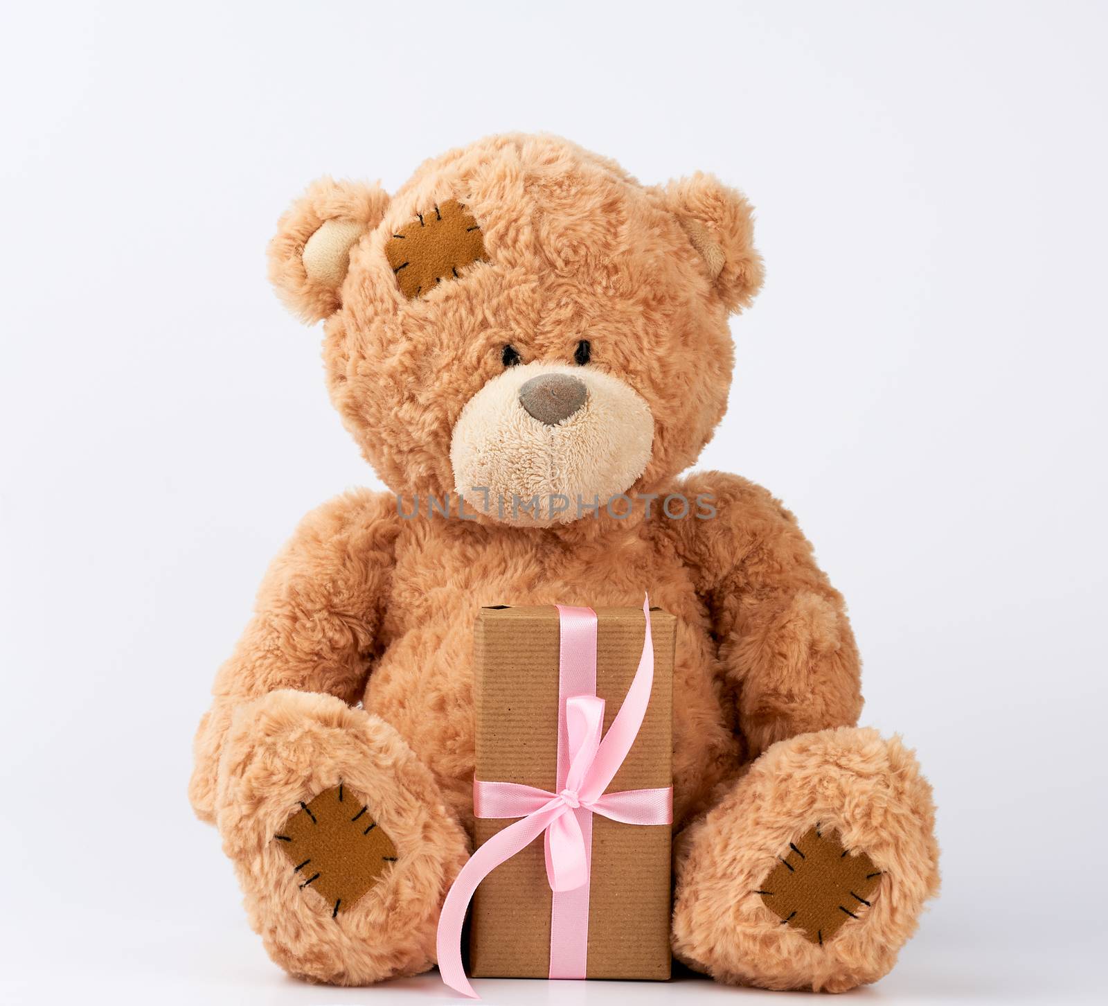 big beige teddy bear with patches hold gift in boxe wrapped in brown paper, tied with pink silk ribbon on a white background, happy birthday and valentines day