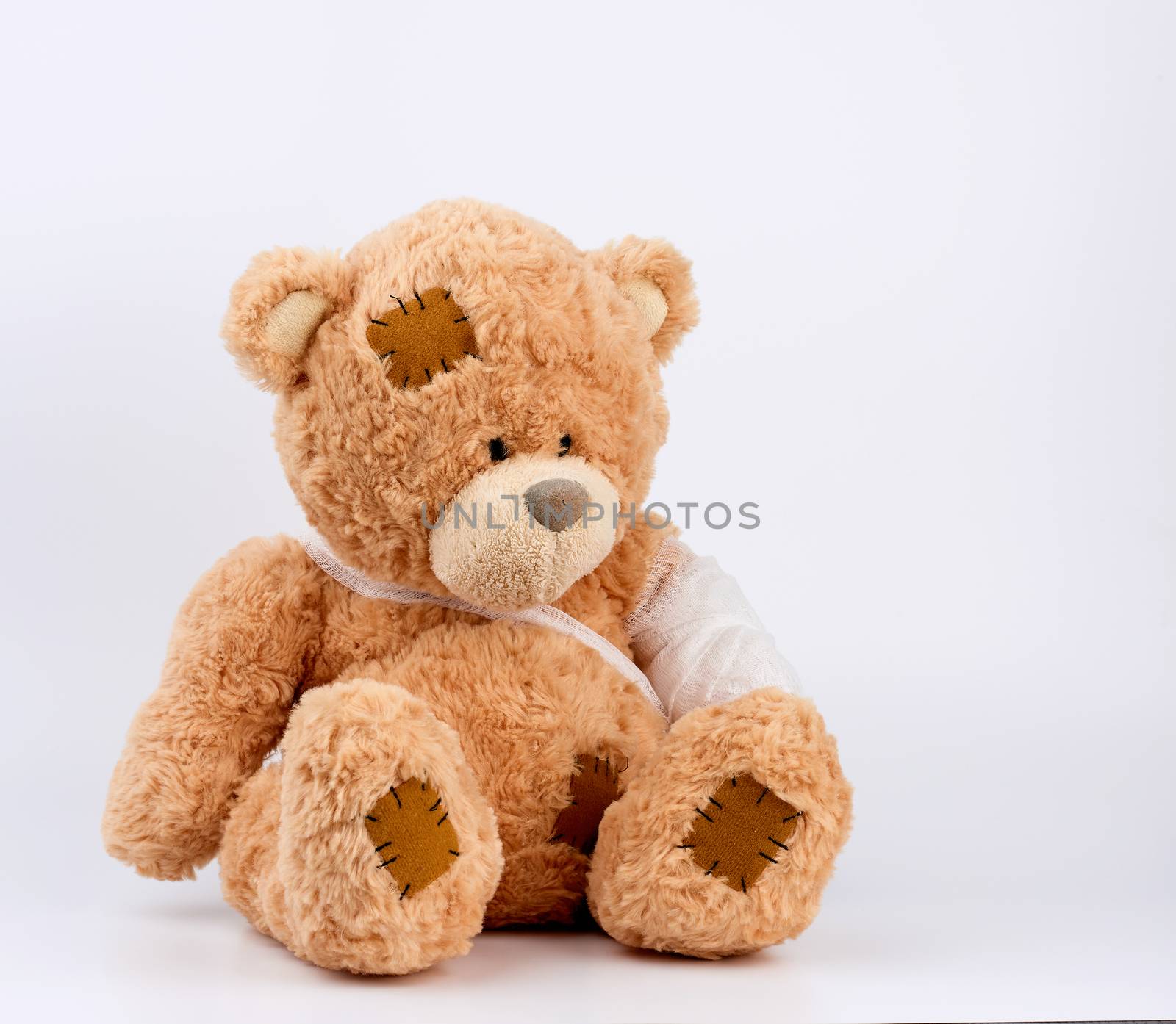 large beige teddy bear with patches sits on a white background, left paw is bandaged with a white medical bandage, concept of pediatrics, treatment of animals