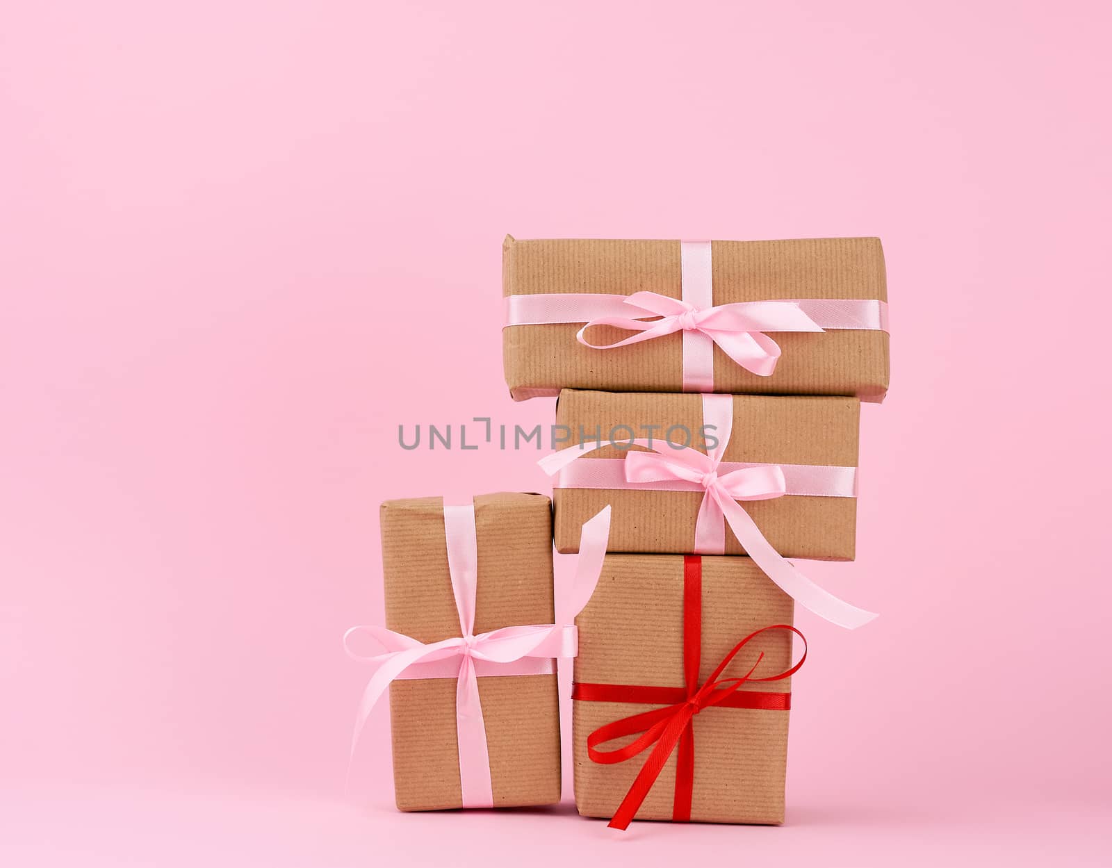 stack of gifts in boxes wrapped in brown kraft paper and tied with silk ribbon on a pink background. Festive concept