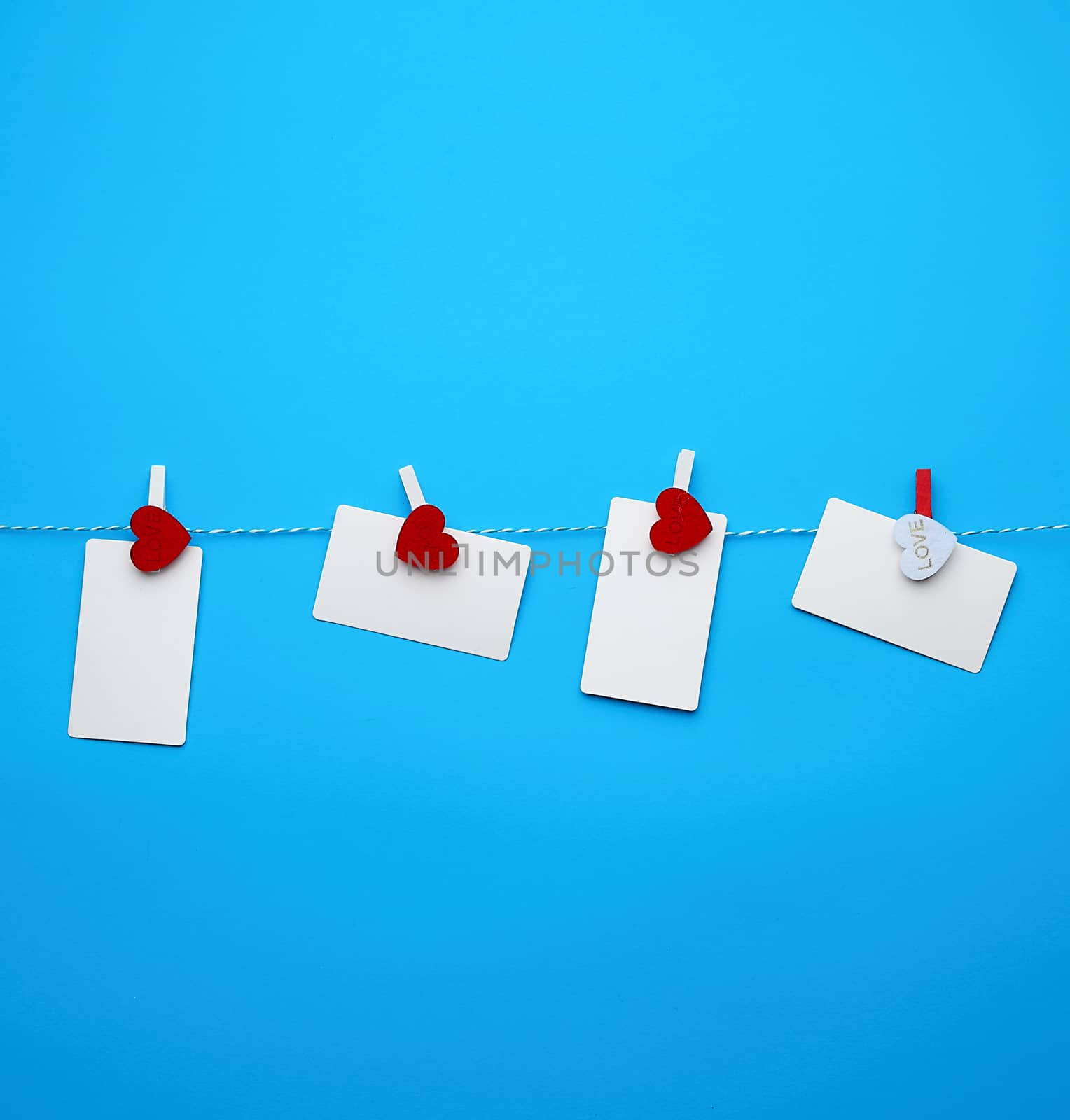 white empty paper rectangular business cards on decorative clothespins with a red heart hanging on a rope, blue background, place for an inscription