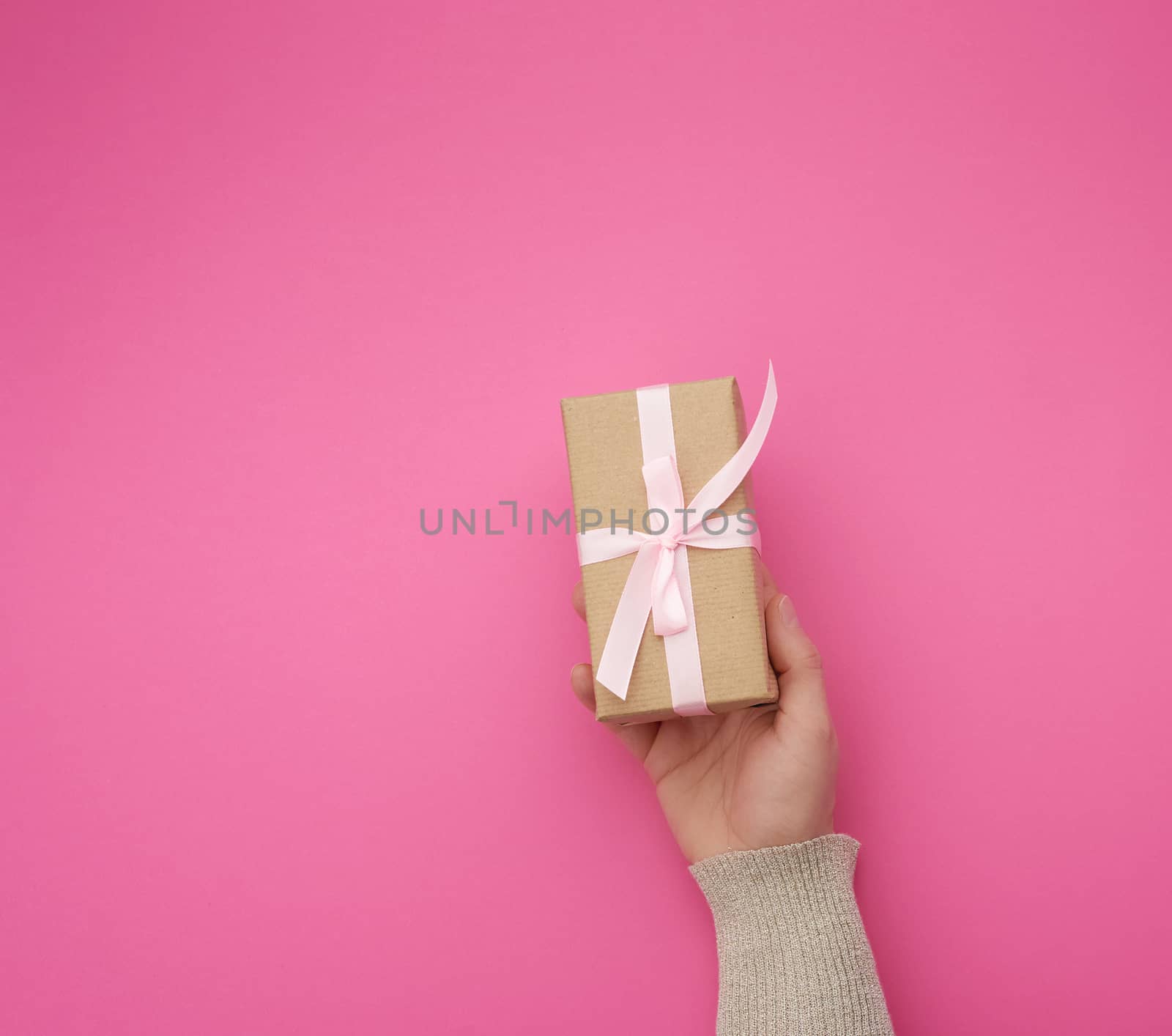 female hand holds a brown box with a pink bow on a pink background, concept of a congratulation on a happy birthday, place for text