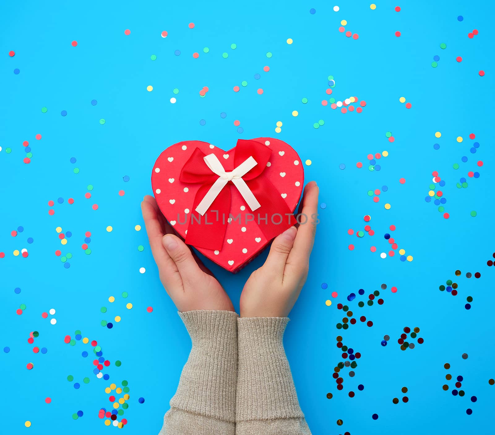 two female hands holding a red cardboard box with a bow on a blue background with multi-colored confetti, concept of giving a gift for a holiday