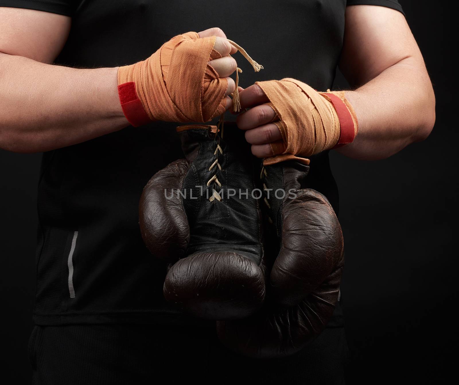 athlete in a black uniform holds very old brown boxing gloves in his hand, his hands are bandaged with an orange elastic sports bandage, black background, copy space