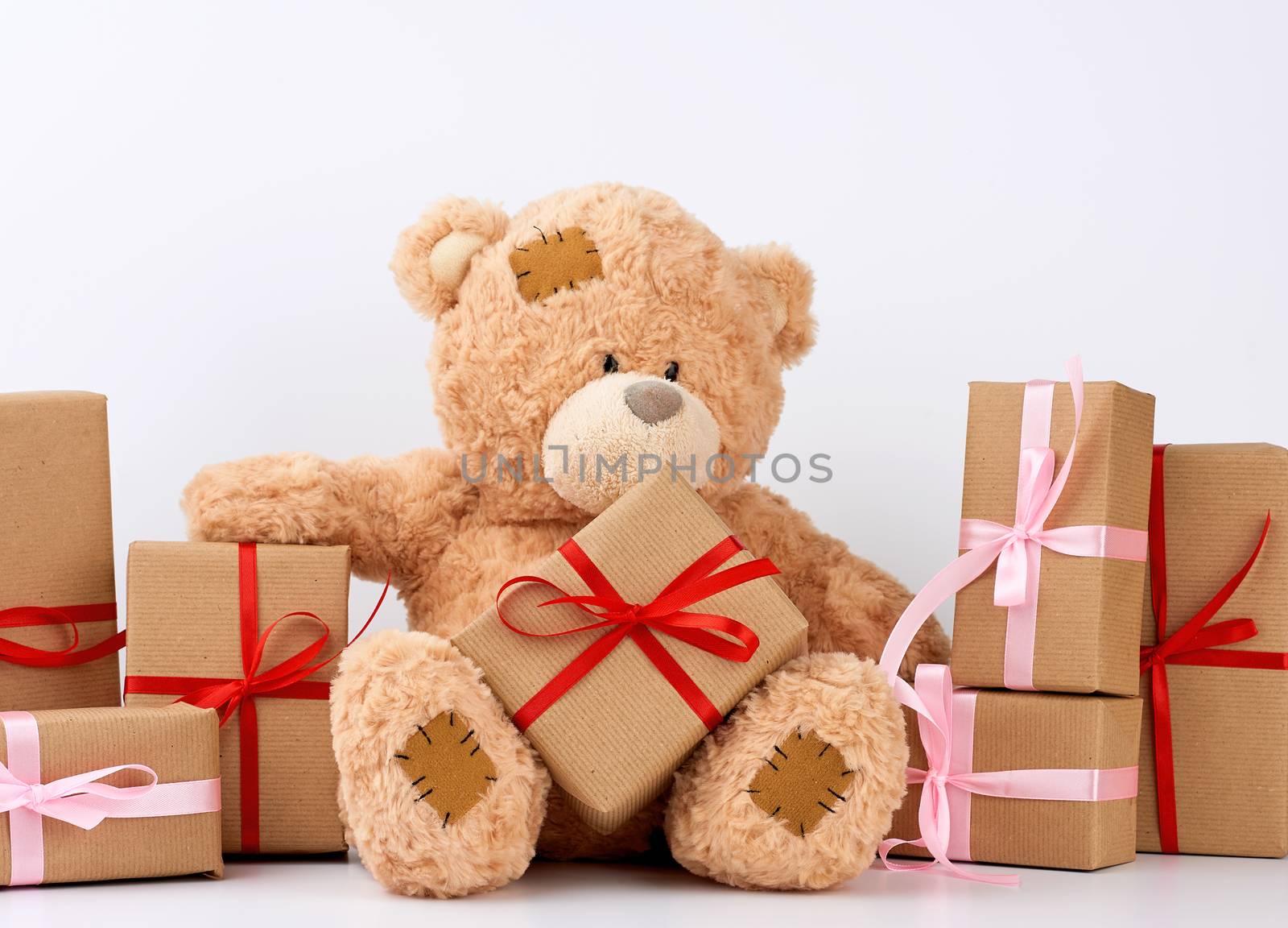big beige teddy bear with patches, a stack of gifts in boxes wrapped in brown paper, tied with silk ribbon on a white background, happy birthday and valentines day