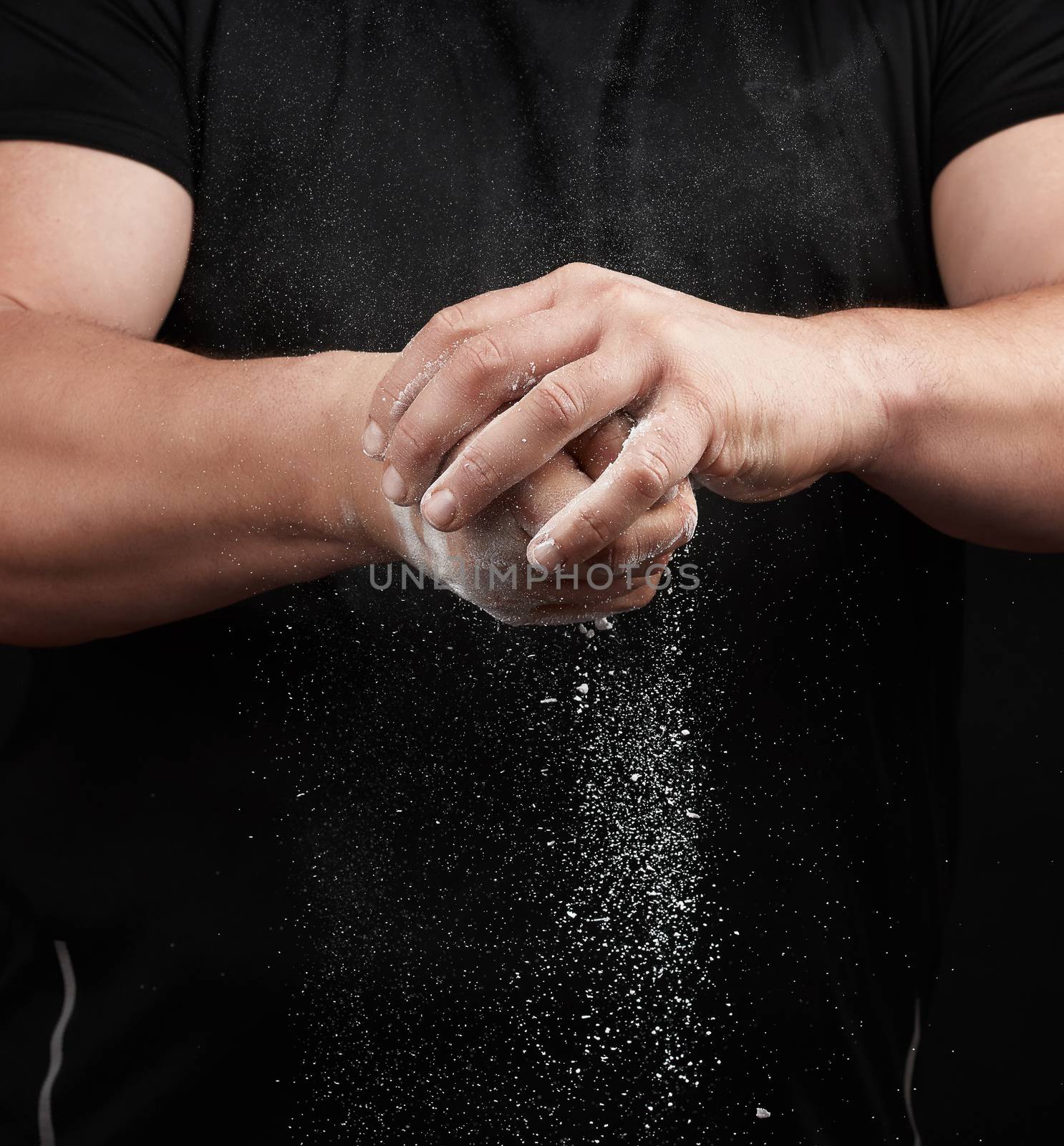 muscular athlete in a black uniform rubs his hands with white dry sports magnesia, powder scatters in different directions on a black background