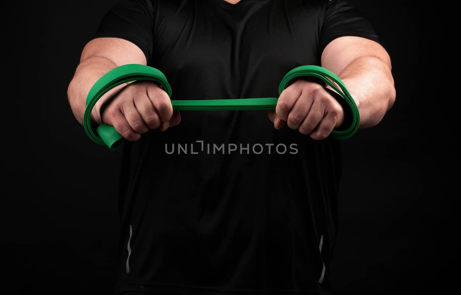 athlete with a muscular body in black clothes is doing physical exercises with green rubber, low key