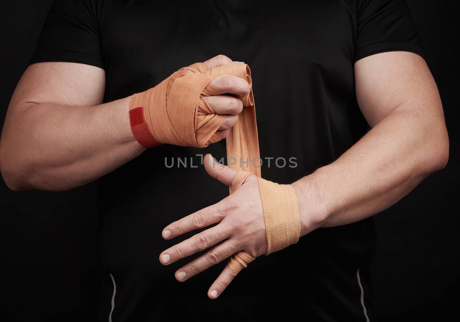 athlete stand in black clothes and wrap his hands in red textile elastic orange bandage before training, black background, muscular body