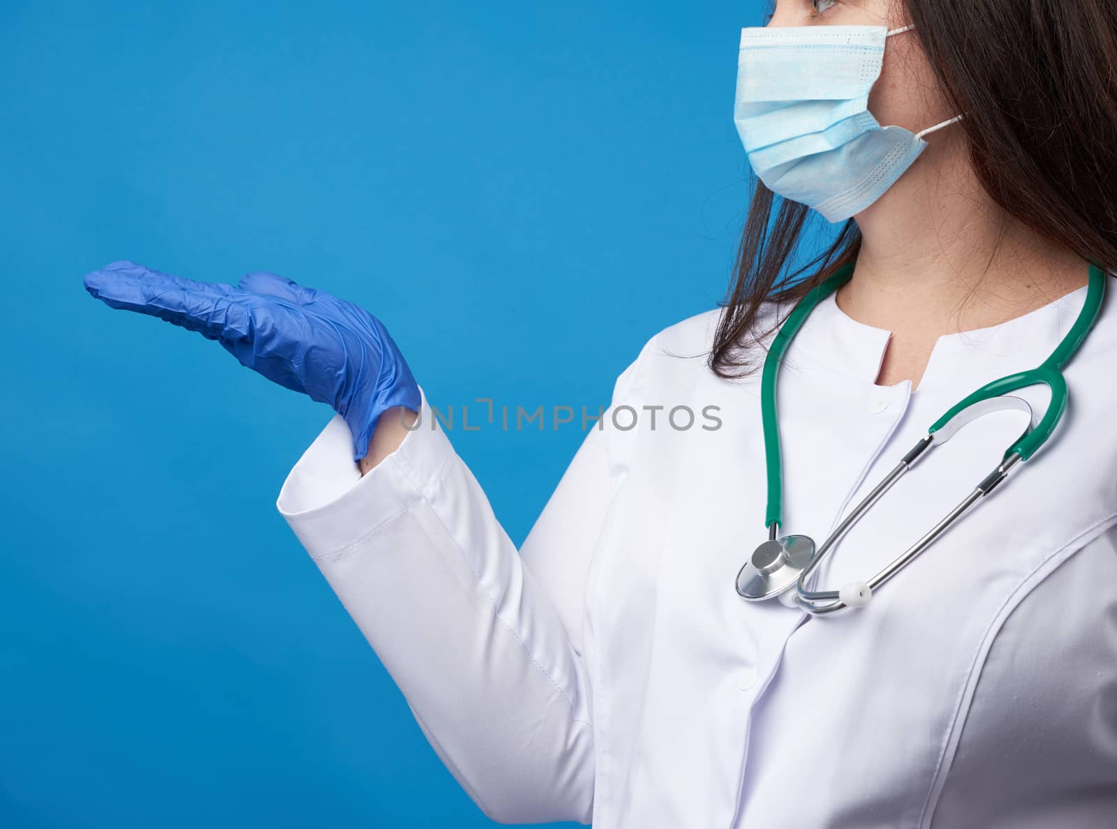 young woman doctor in a white coat, blue latex gloves and a protective mask on his face conditionally holds an object with his right hand on a blue background
