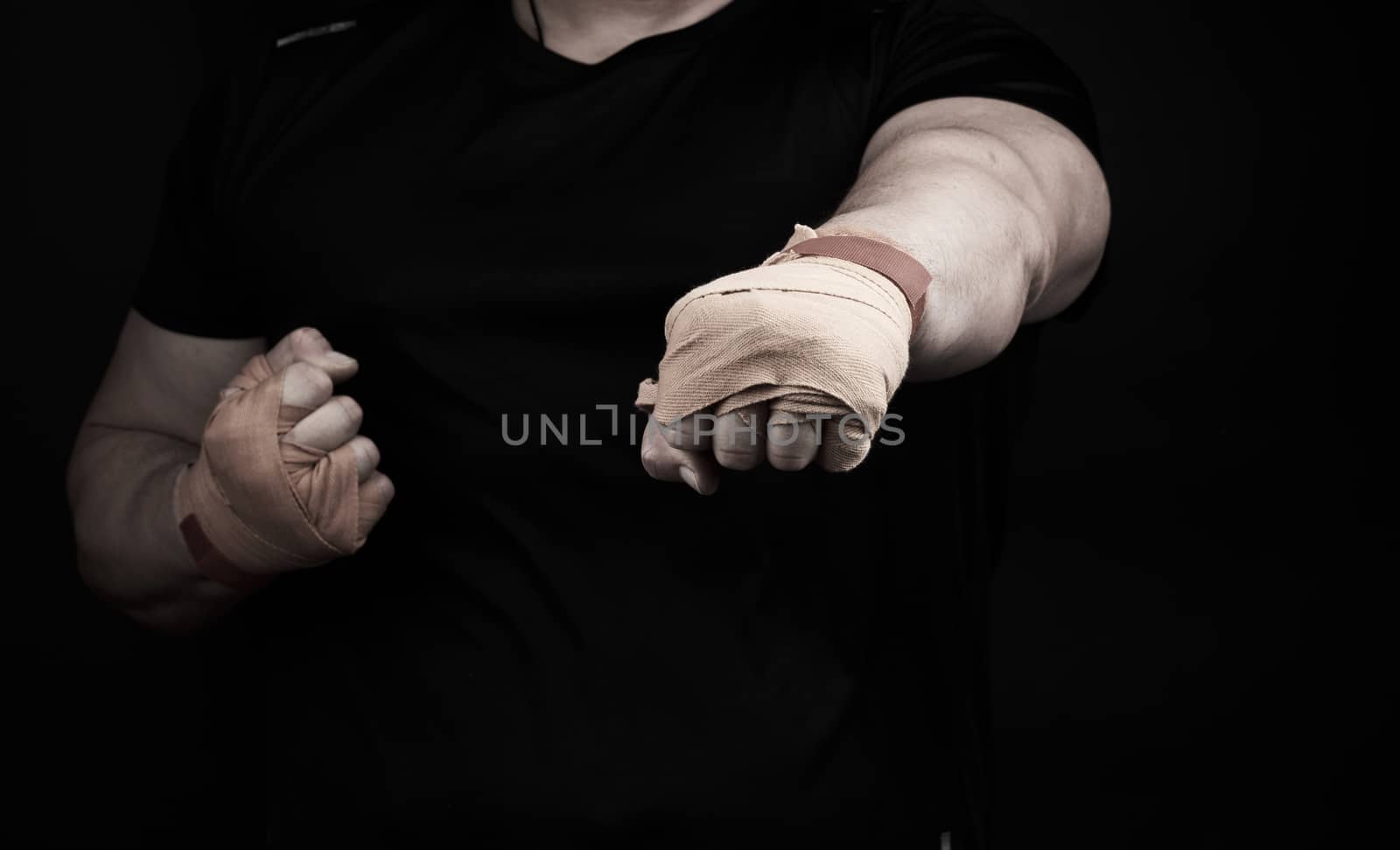 adult man in a black uniform and muscular arms stands in a sports stance, holds a sports elastic bandage on his hands, punch forward, dark background