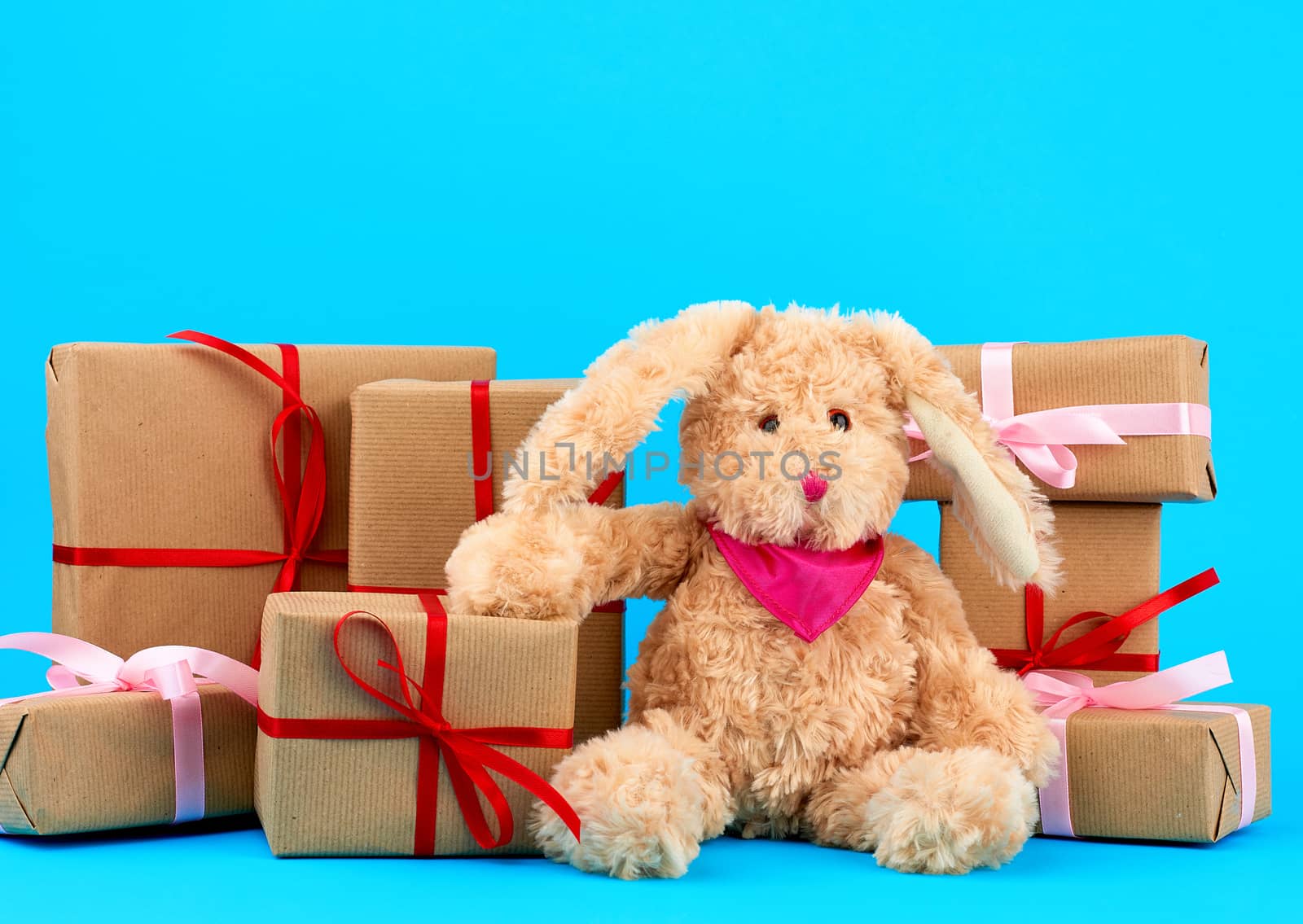 cute little brown plush rabbit and a stack of boxes wrapped in brown kraft paper and tied with silk ribbon on a blue background, concept of holiday, easter