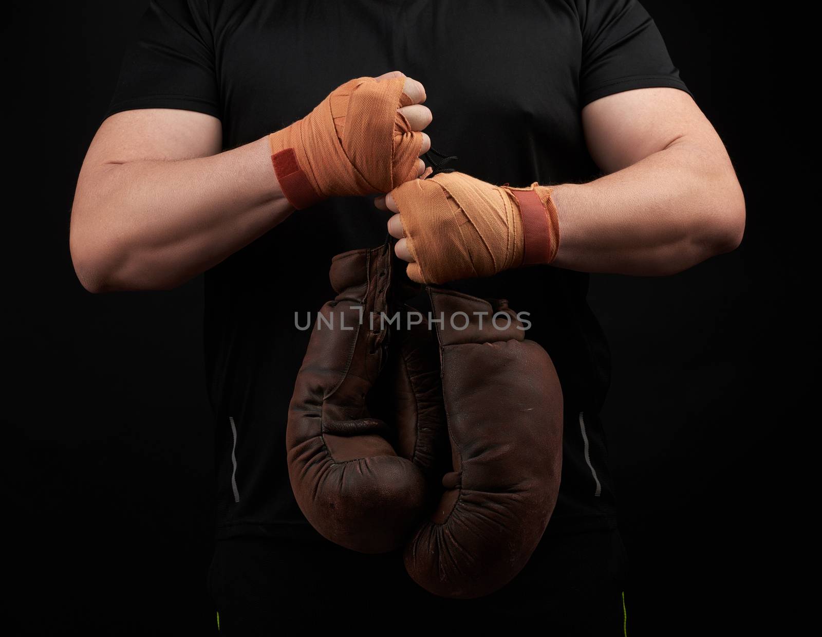 muscular athlete in a black uniform holds very old brown boxing gloves in his hand, his hands are bandaged with an orange elastic sports bandage, black background