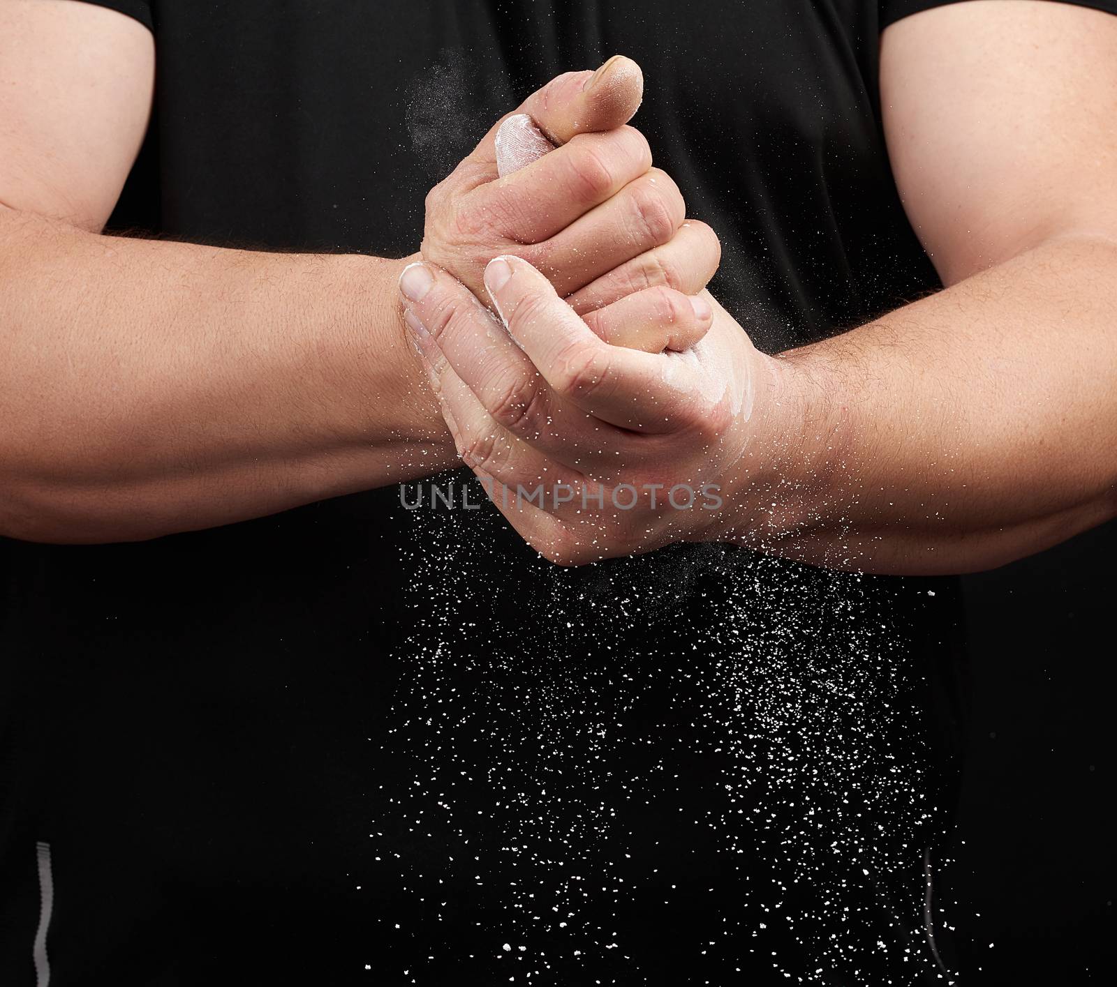 muscular athlete in a black uniform rubs his hands with white dry sports magnesia, powder scatters in different directions on a black background