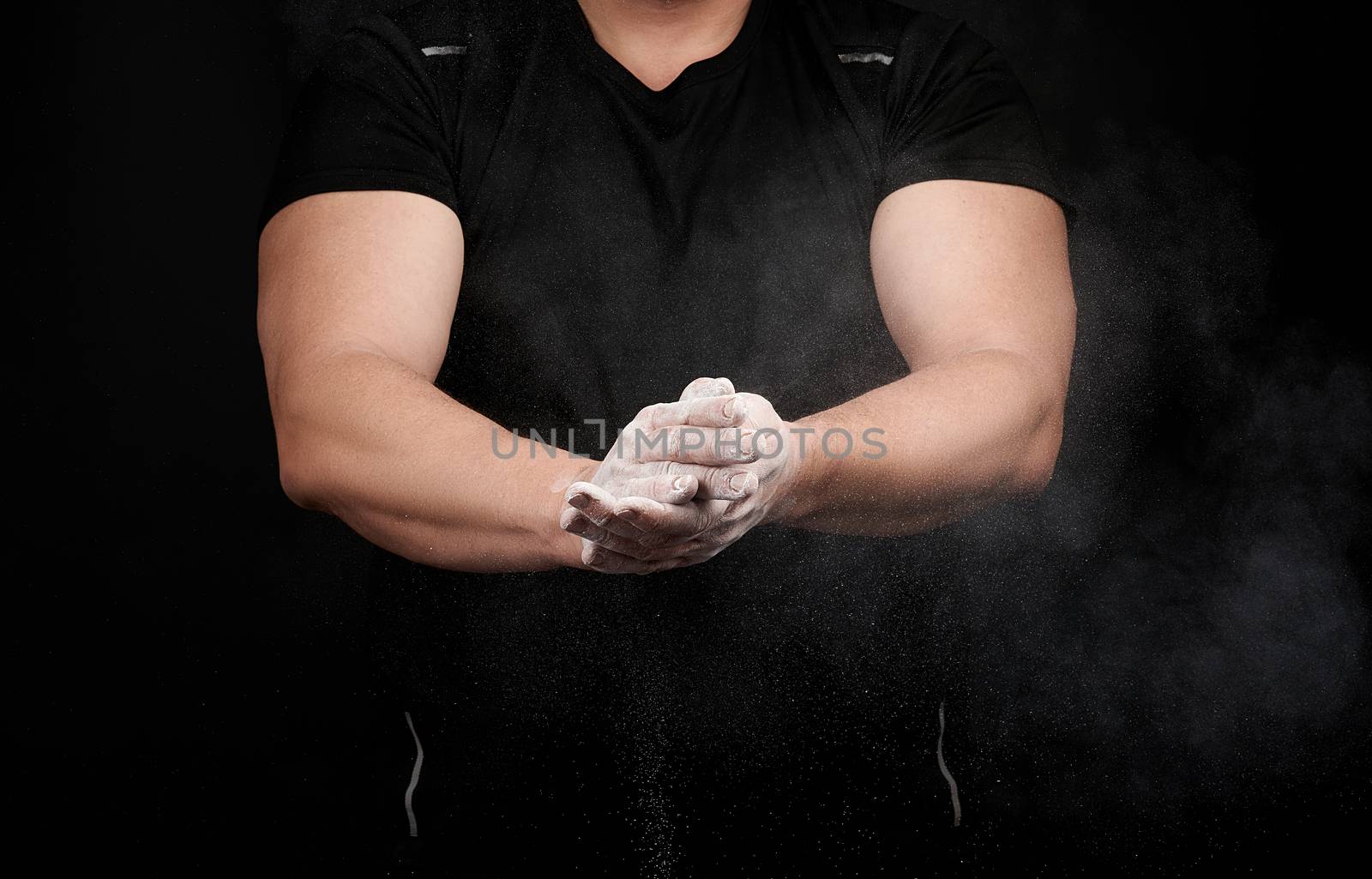 muscular athlete in a black uniform rubs his hands with white dry sports magnesia, powder scatters in different directions on a black background