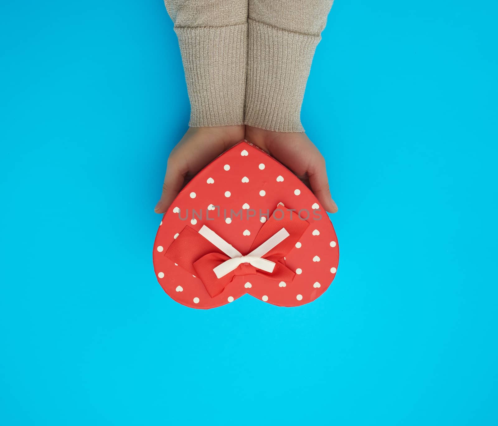 female hand holding a red cardboard box with a bow on a blue background, top view