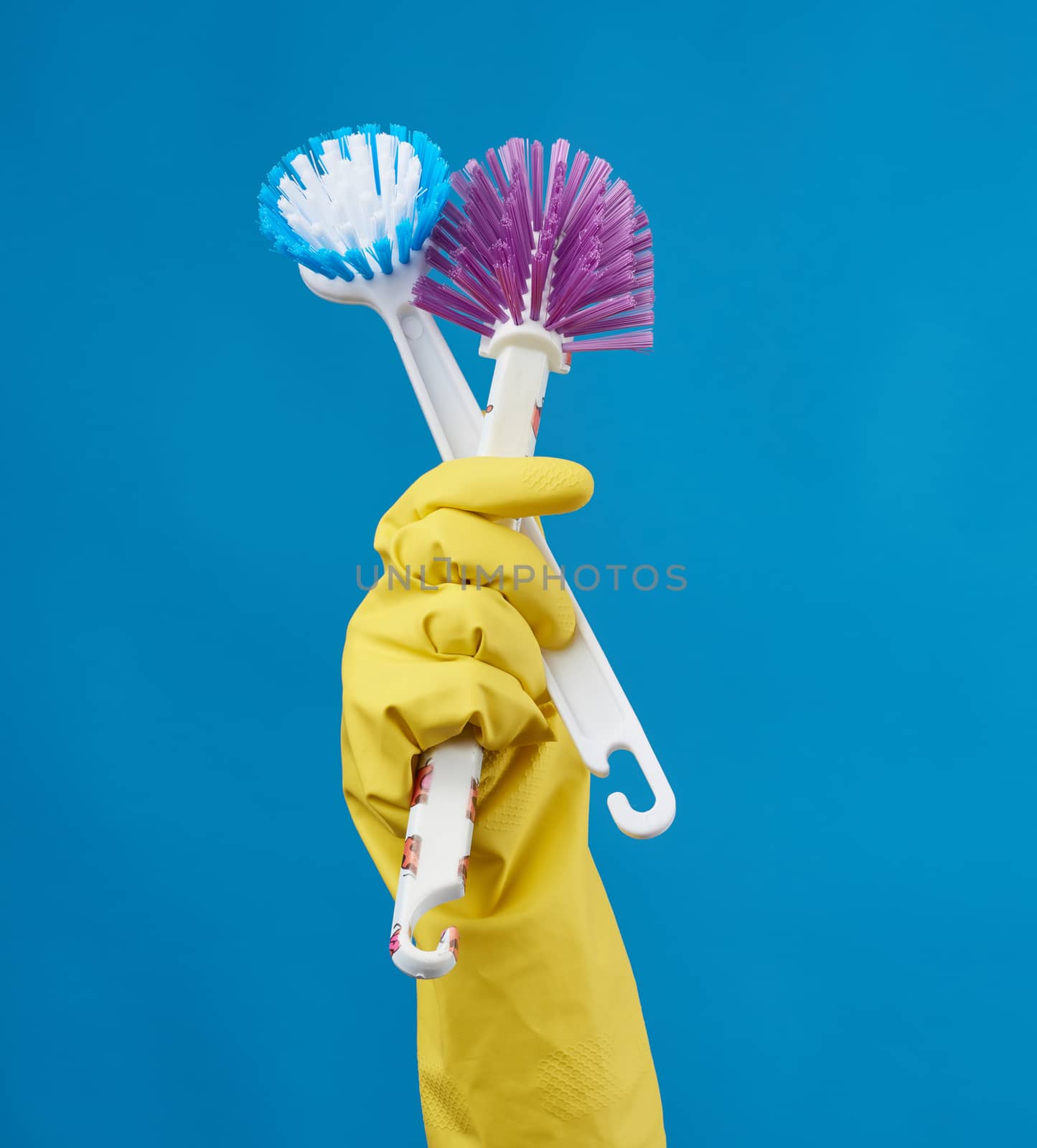 hand in a yellow rubber glove for cleaning a house holds two plastic brushes with a handle on a blue background