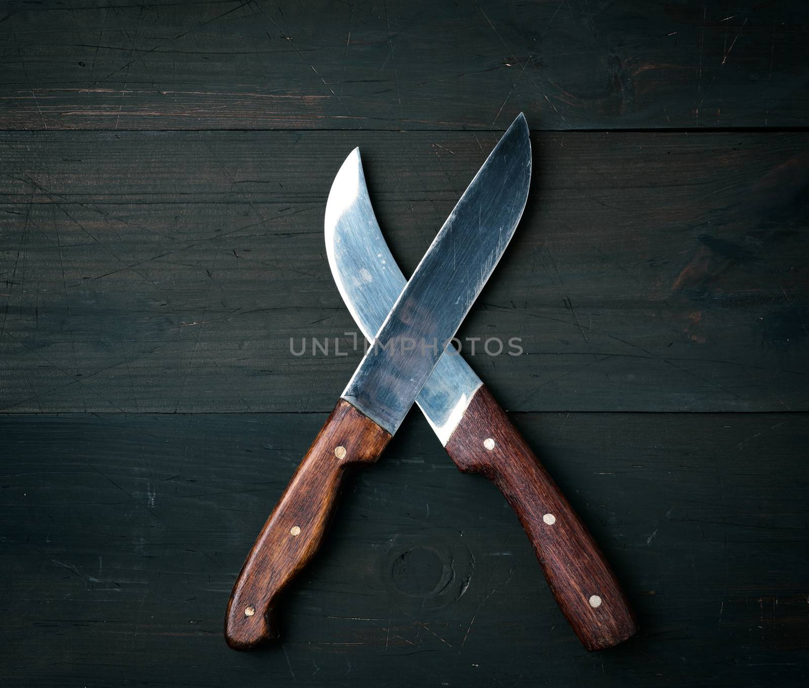 two sharp knives lie on a brown wooden surface, kitchen items are crossed, top view