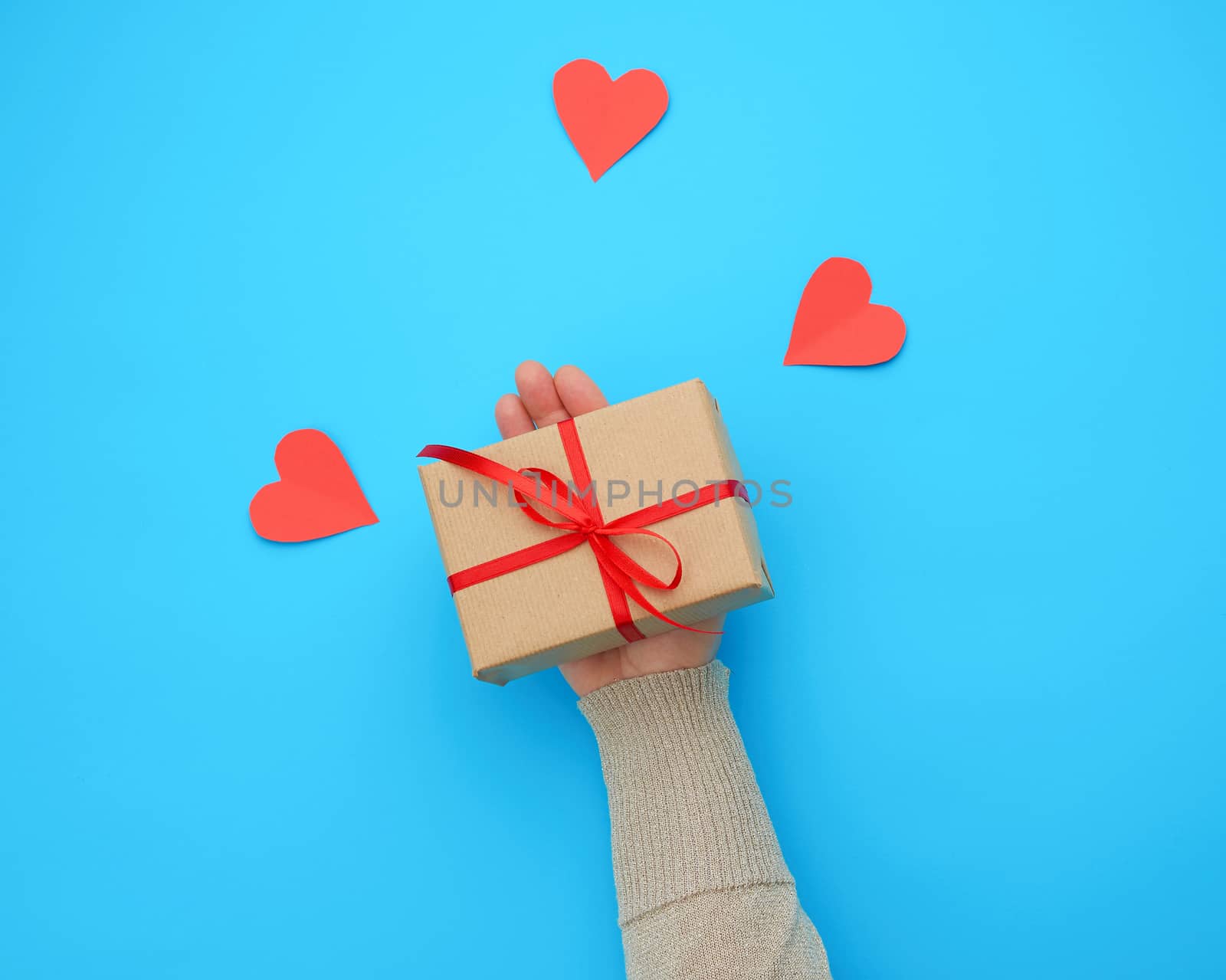 female hand holds a box wrapped in brown kraft paper and tied with a silk ribbon on a blue background, paper red hearts. Valentine's Day backdrop