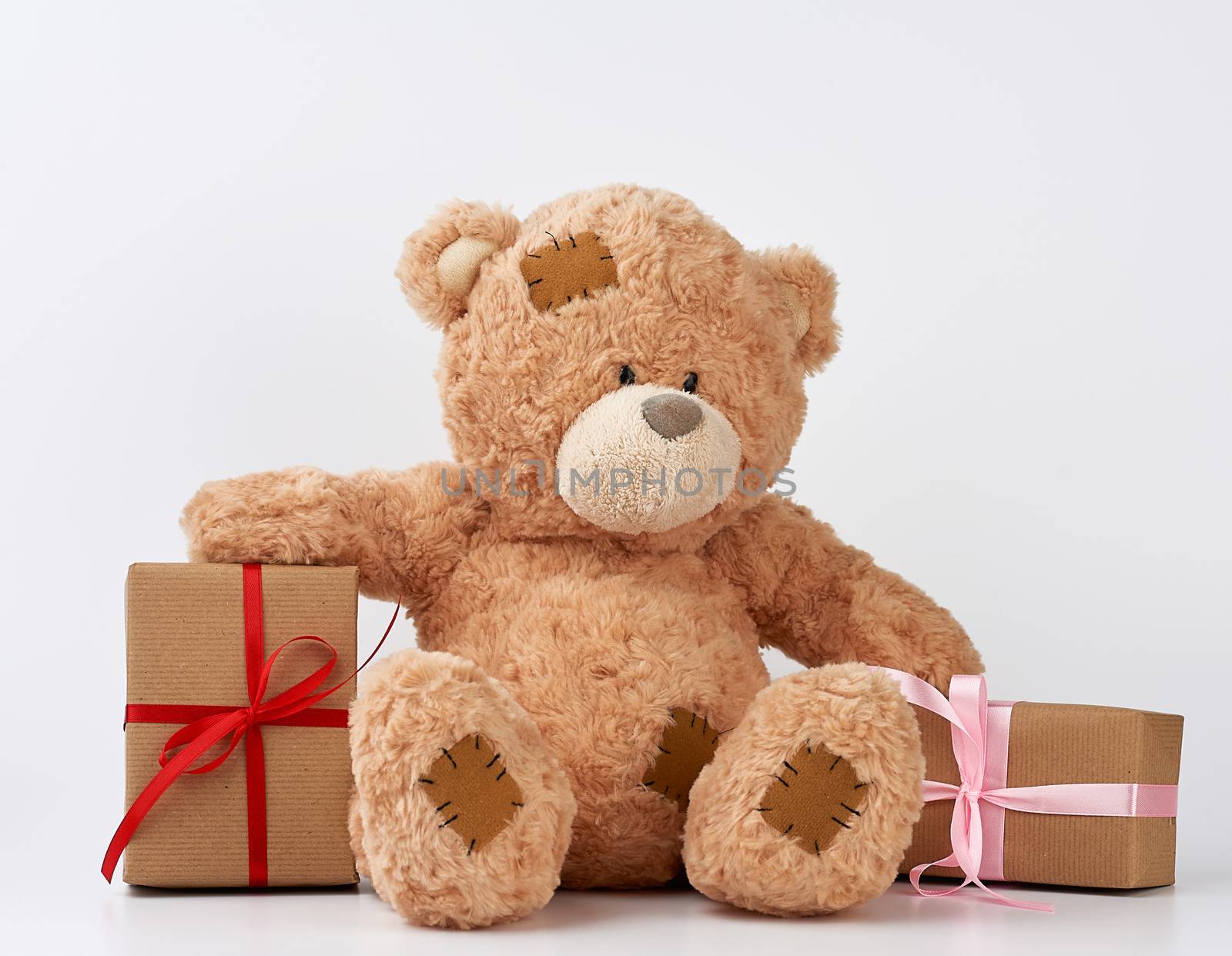 big beige teddy bear with patches, a stack of gifts in boxes wrapped in brown paper, tied with silk ribbon on a white background, happy birthday and valentines day