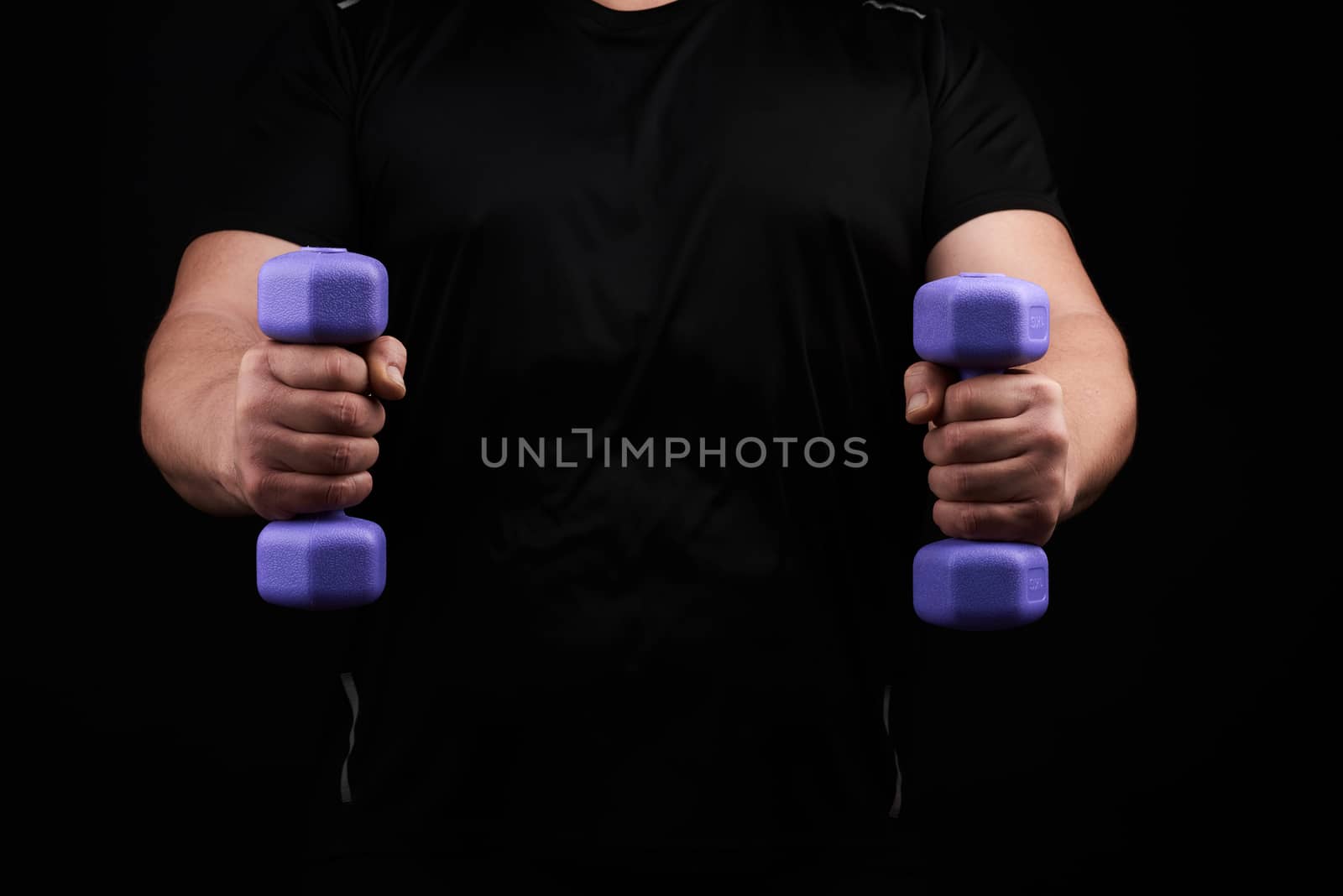 adult male athlete in black uniform holds blue plasti dumbbells in hands, black background