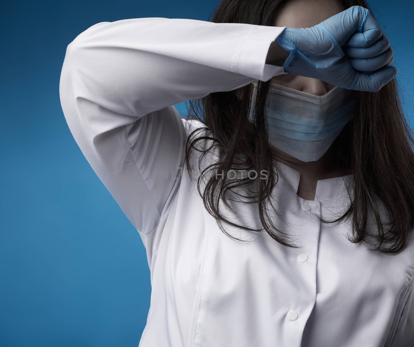young woman doctor in a white coat, blue latex gloves and a protective mask on his face stands on a blue background. Hand pressed to forehead, concept of fatigue and hopelessness.