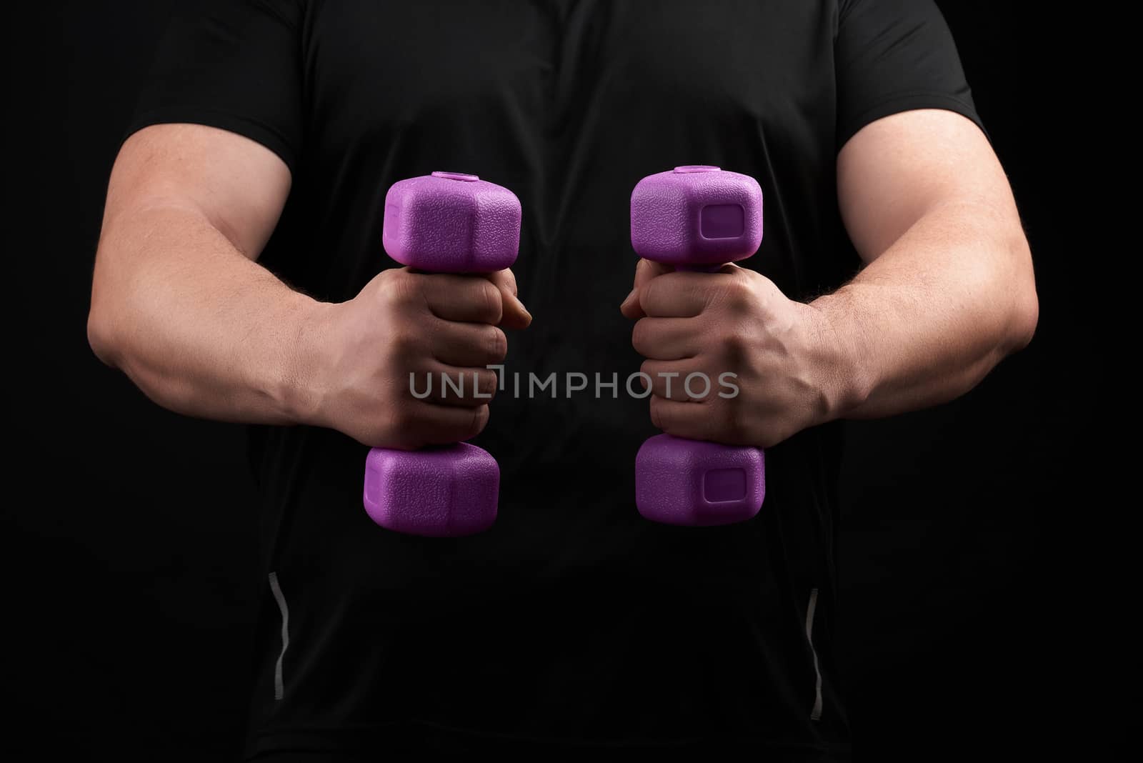 adult male athlete in black uniform holds blue plastic purple dumbbells in hands, black background