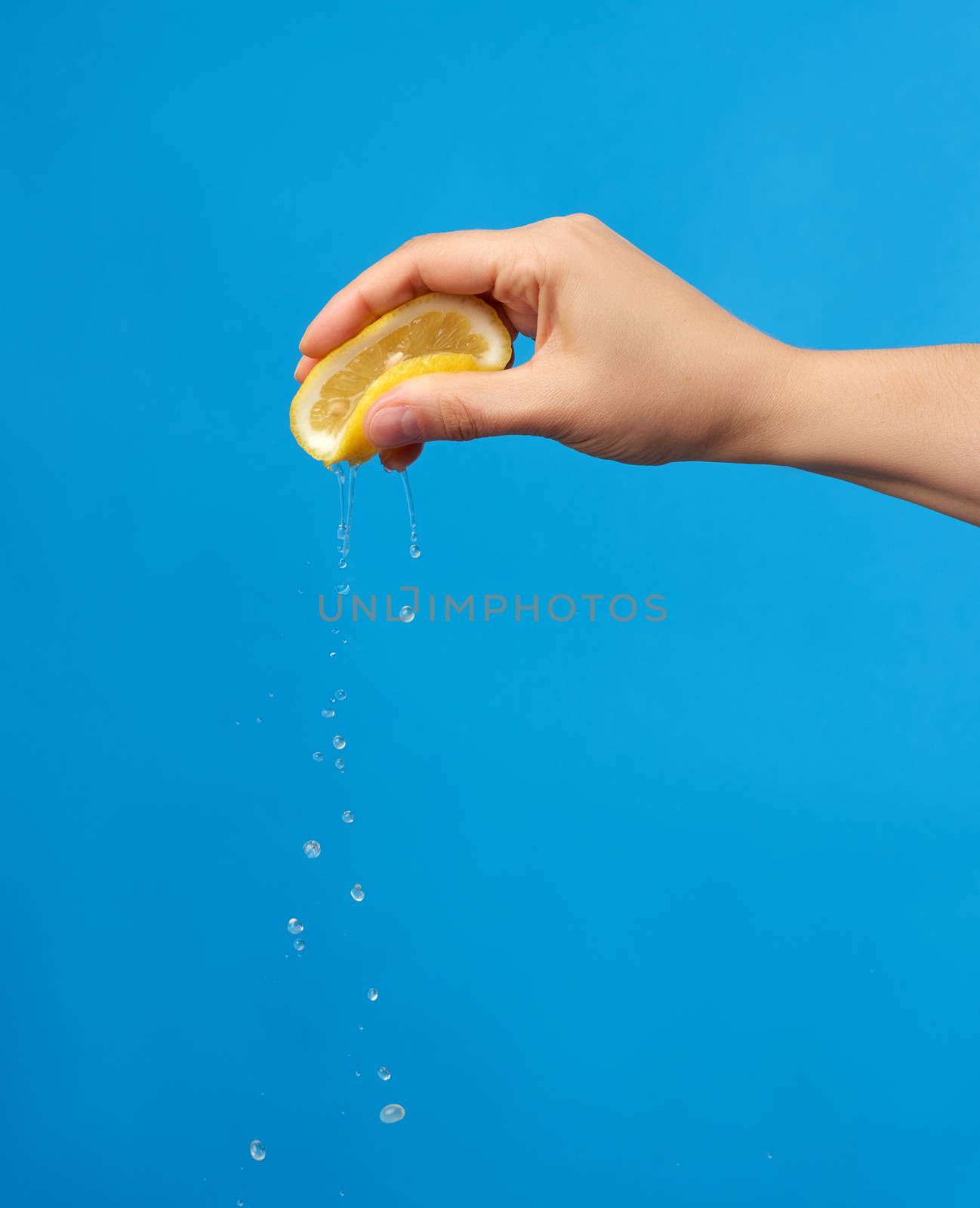 female hand holds half a yellow lemon and squeezes it on a blue  by ndanko