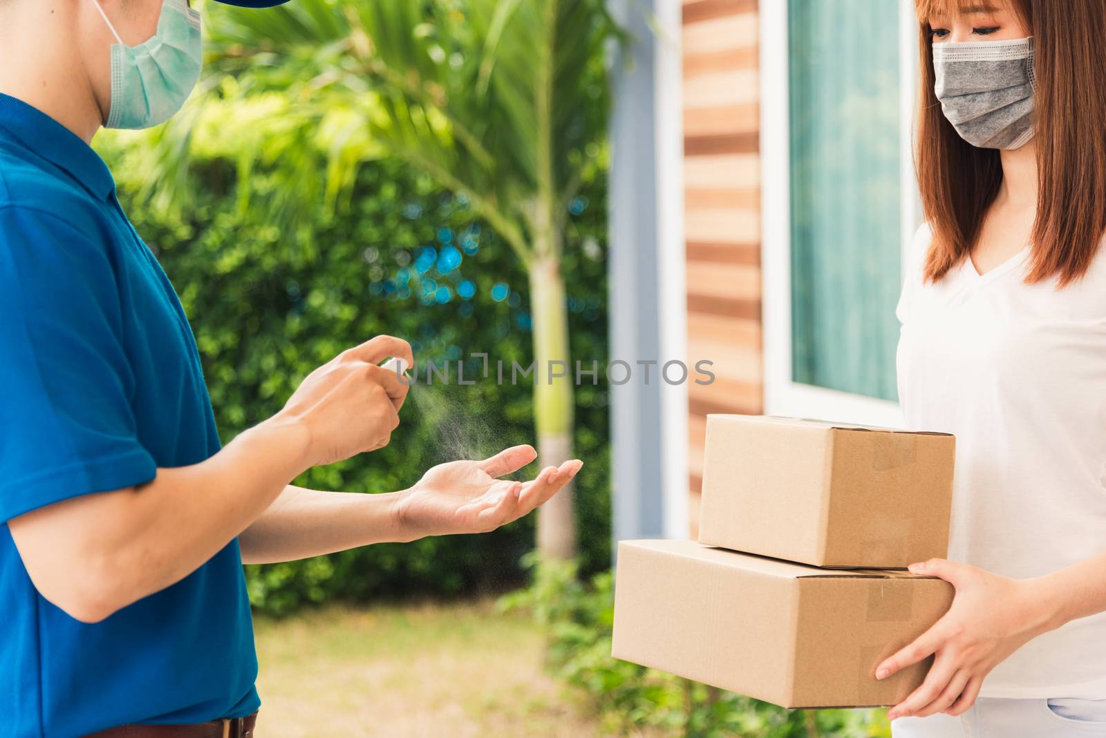 Asian delivery express courier young man use spray sterilize before giving boxes to woman customer receiving both protective face mask, under curfew quarantine pandemic coronavirus COVID-19