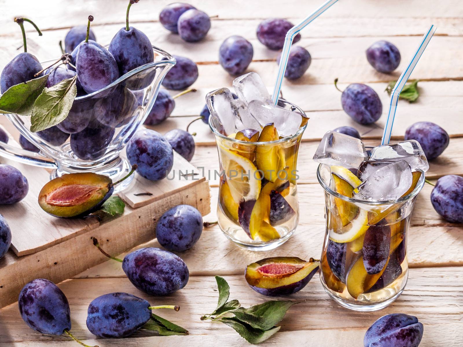 Refreshing drink from fresh juicy sweet sips with lemon in glass mugs with ice cubes on a wooden background with plums and green leaves with plums in a glass sauceboat