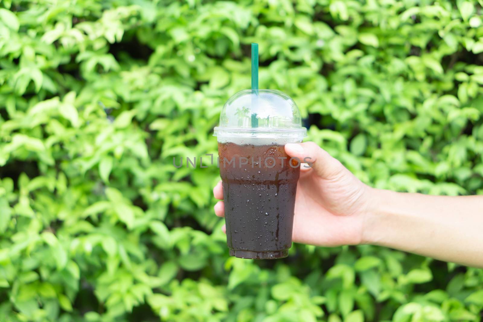 Close up hand holding glass of ice americano coffee with green leaves nature background, selective focus
