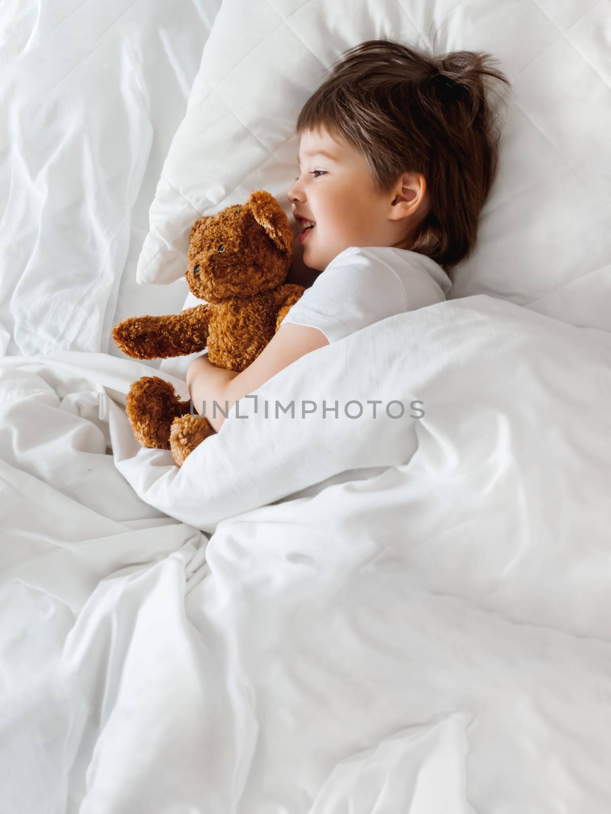 Toddler lies in bed with cute teddy bear. Little boy under white by aksenovko