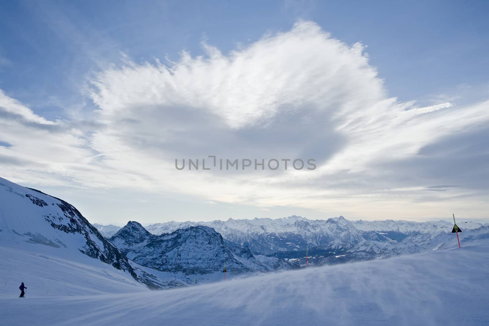 A heart shaped cloud floats over a ski run as part of an expansive mountain vista in the italian alps