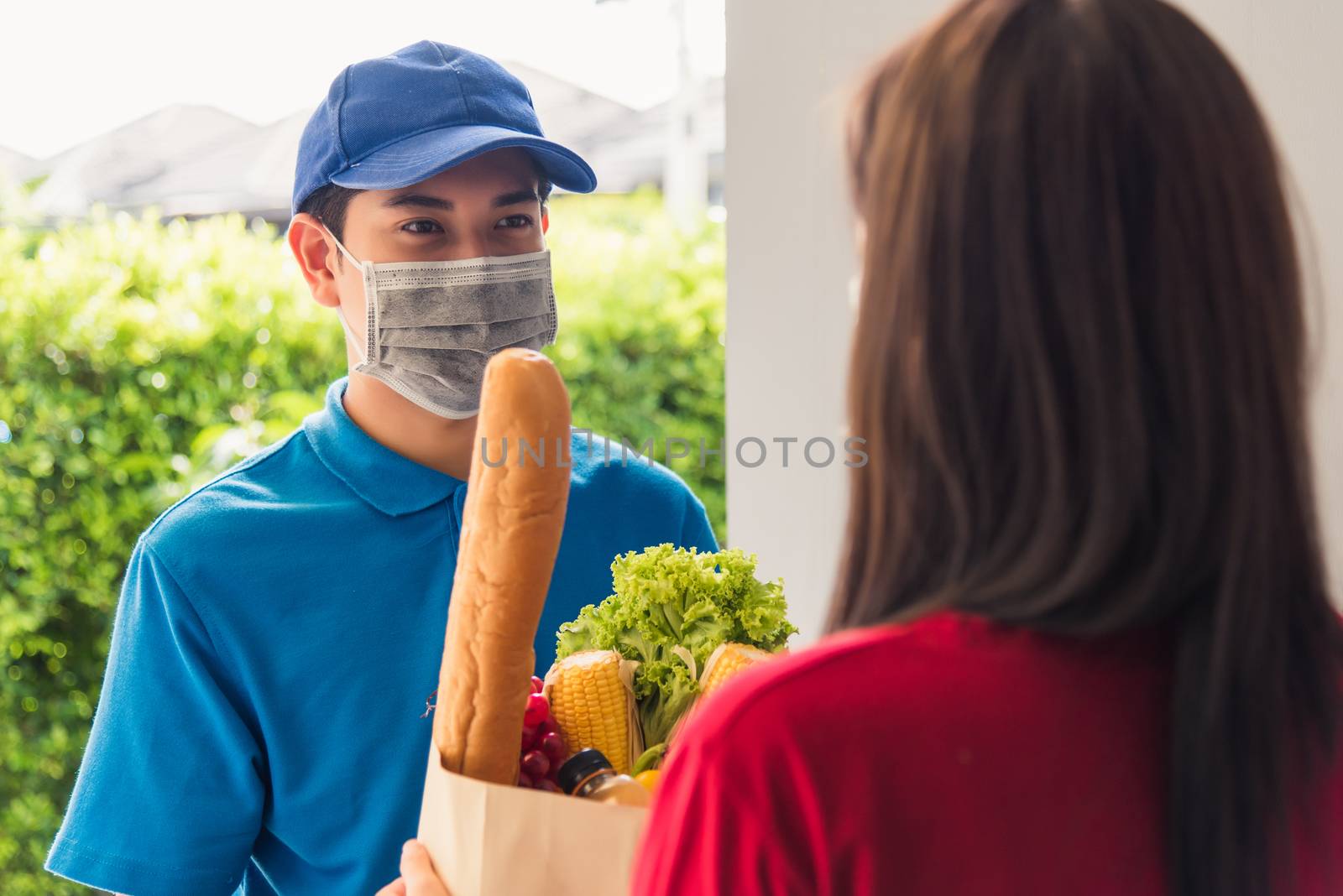 Asian young delivery man in uniform wear protective face mask he making grocery service giving fresh food to woman customer receiving front house under pandemic coronavirus, Back to new normal concept