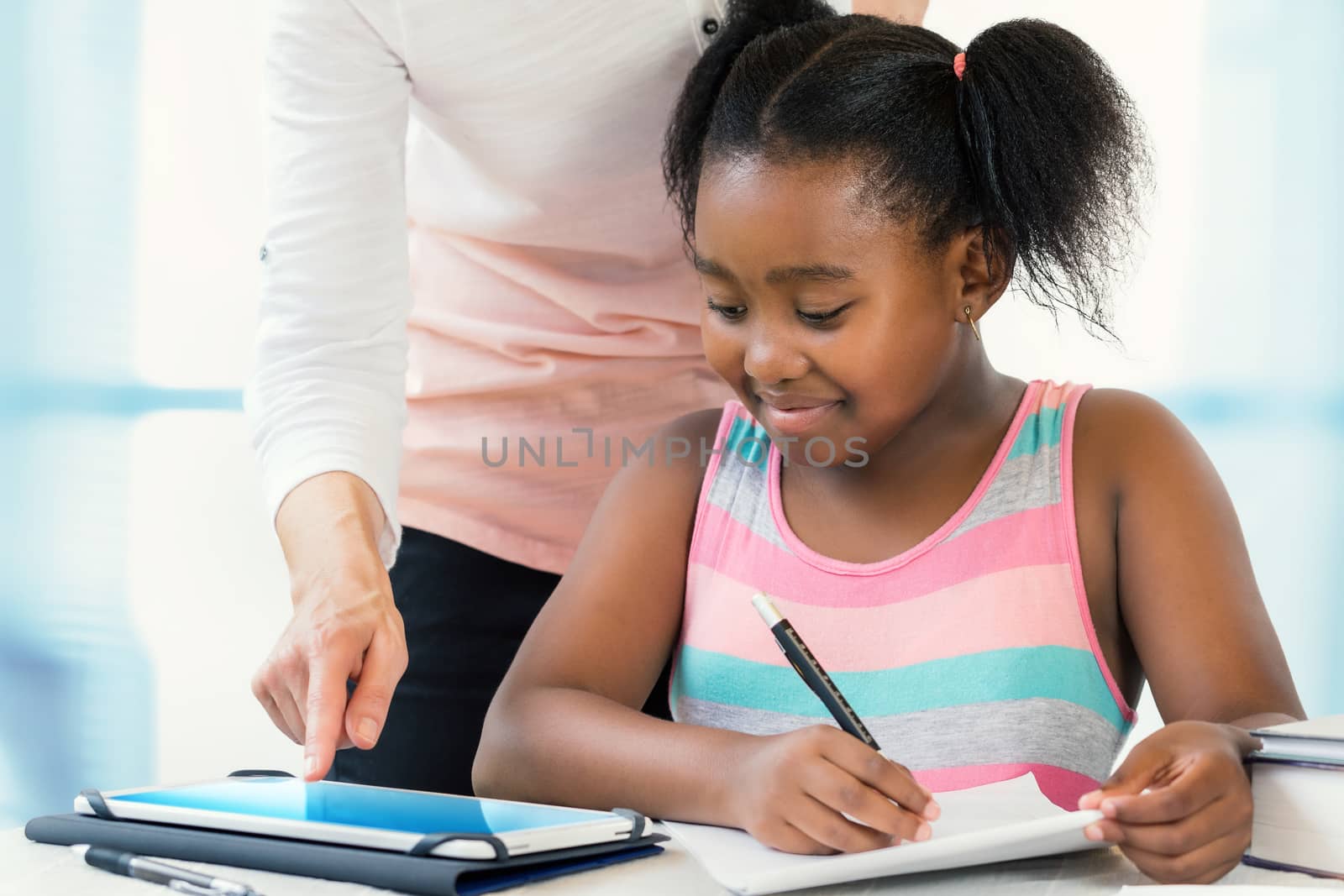 Close up portrait of little african girl struggling with school work.Caucasian teacher showing results on digital tablet at desk.
