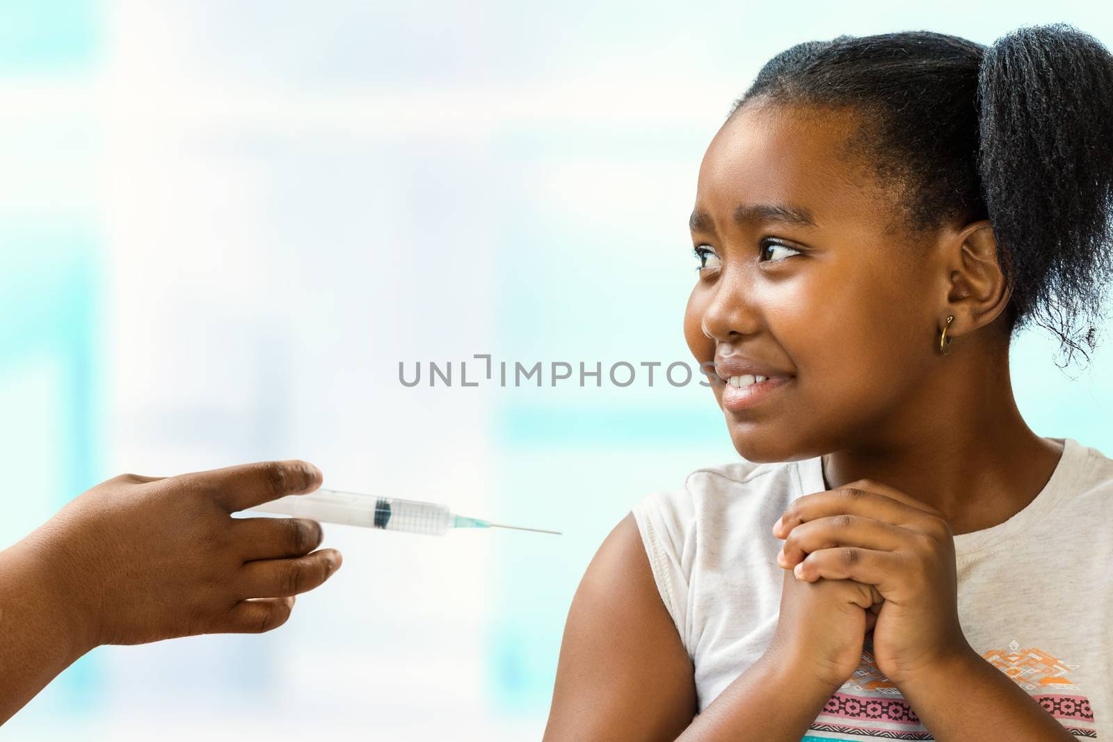 Close up portrait of anxious little african girl scared of syringe with needle.Kid looking aside at doctor with nervous facial expression. Hand holding syringe near upper arm.