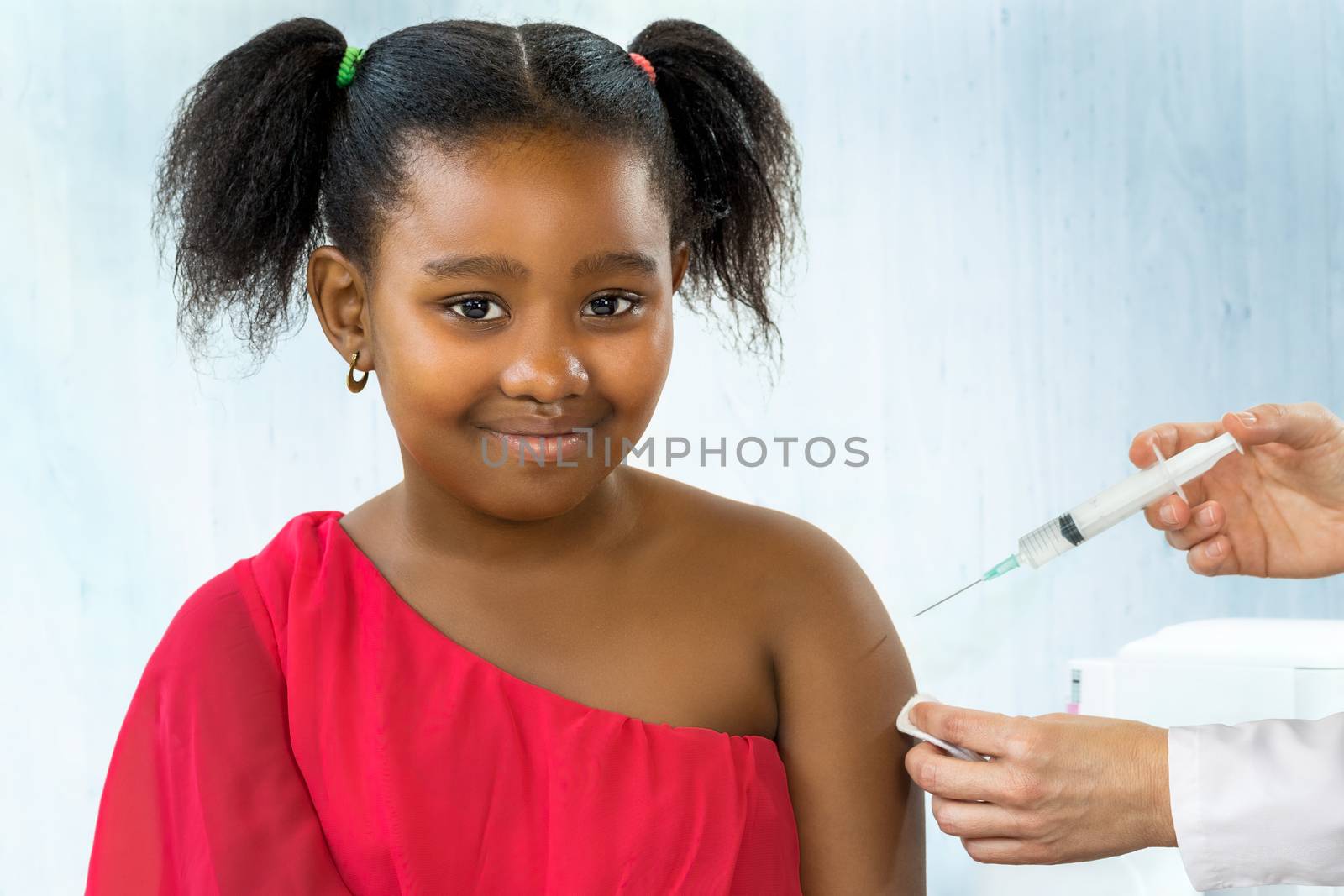 Close up portrait of cute ponytailed african girl receiving disease prevention vaccine.Caucasian hands holding syringe with needle next to upper arm.