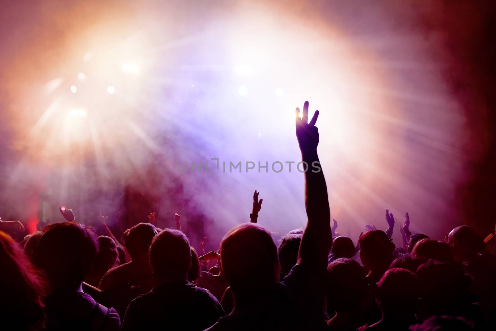 Dancing Crowd of young people dancing at disco. People Raising hands against purple and pink smoky background with light beams.