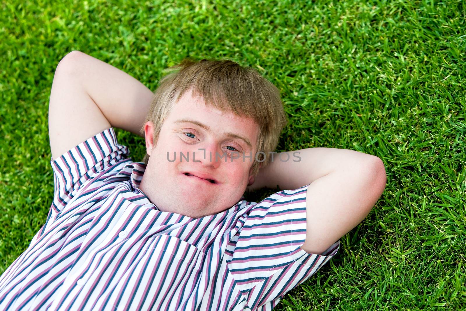 Close up portrait of handicapped boy laying with hands under head on green grass.