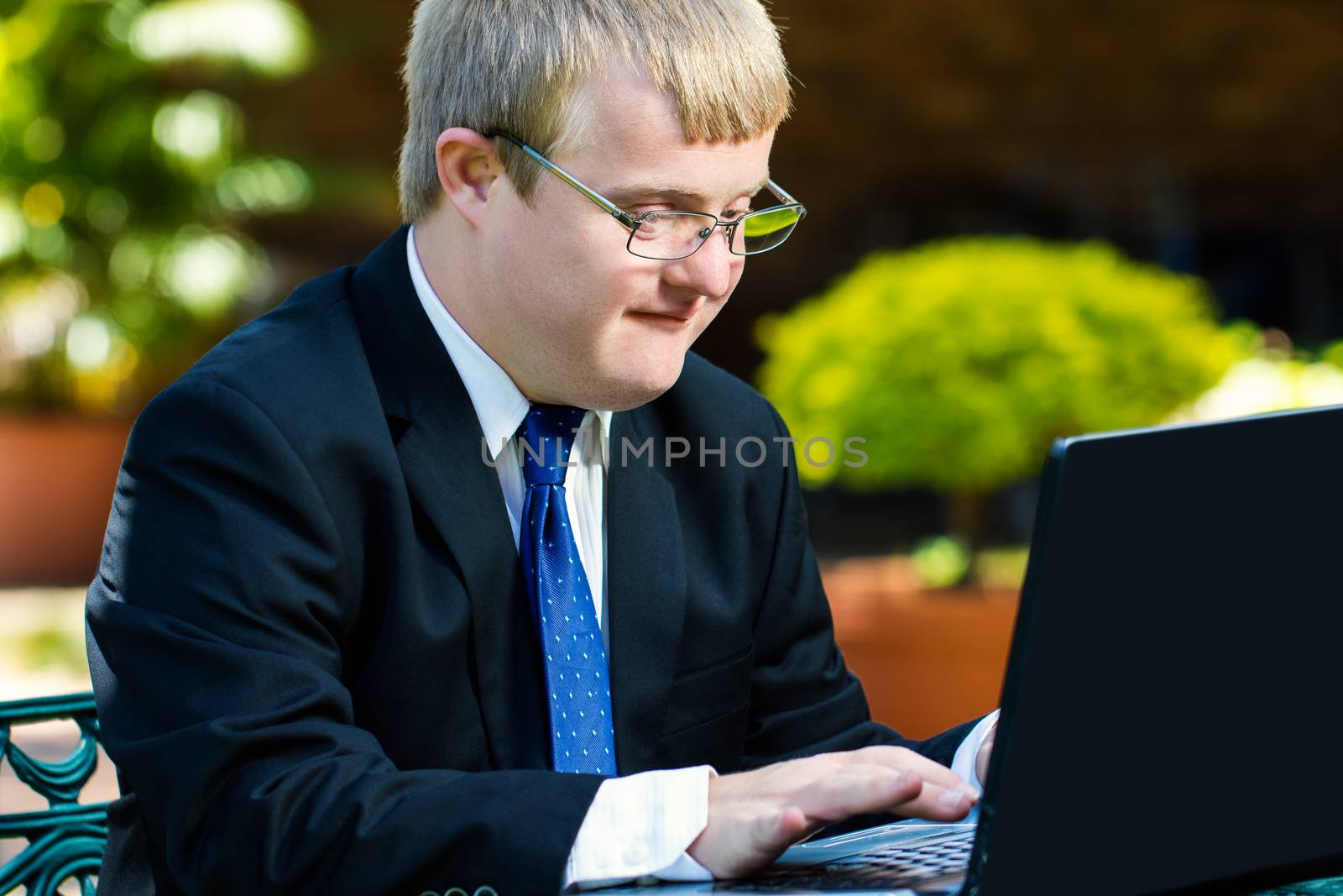 Close up portrait of businessman with down syndrome working. Young man in suit working on laptop in garden.