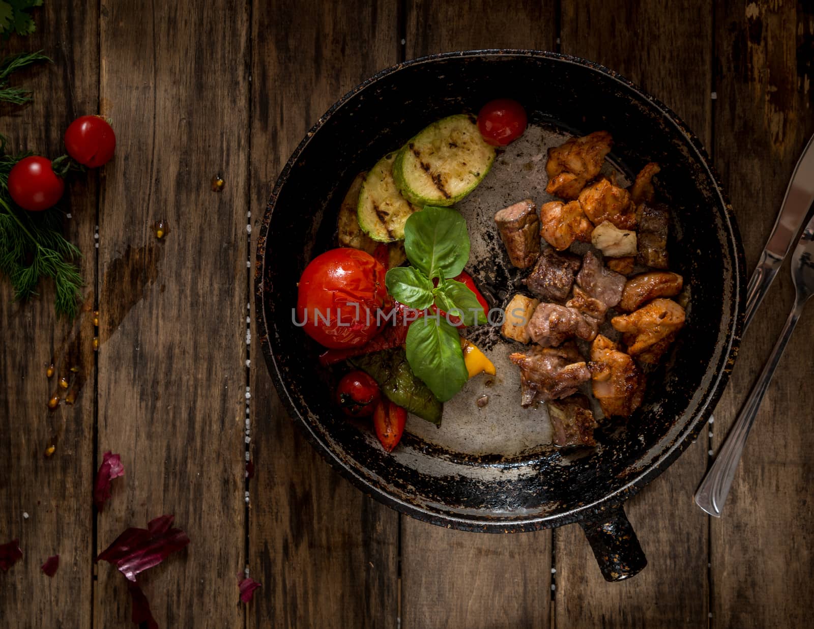 fried pieces of meat and vegetables on an old frying pan, view from above. wooden background