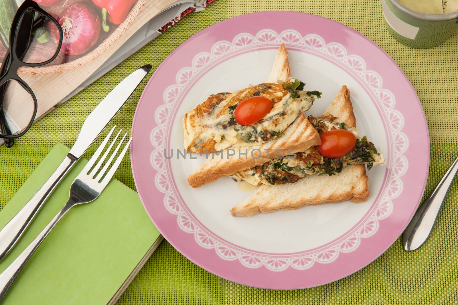 sandwiches with tomatoes in a plate with book and glasses and cutlery on a green tablecloth