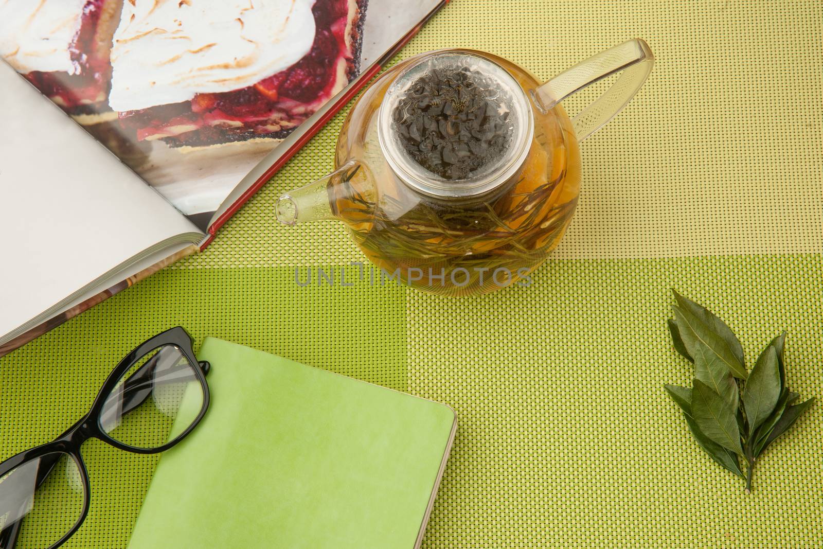 Transparent teapot with green tea with book and glasses on a green tablecloth