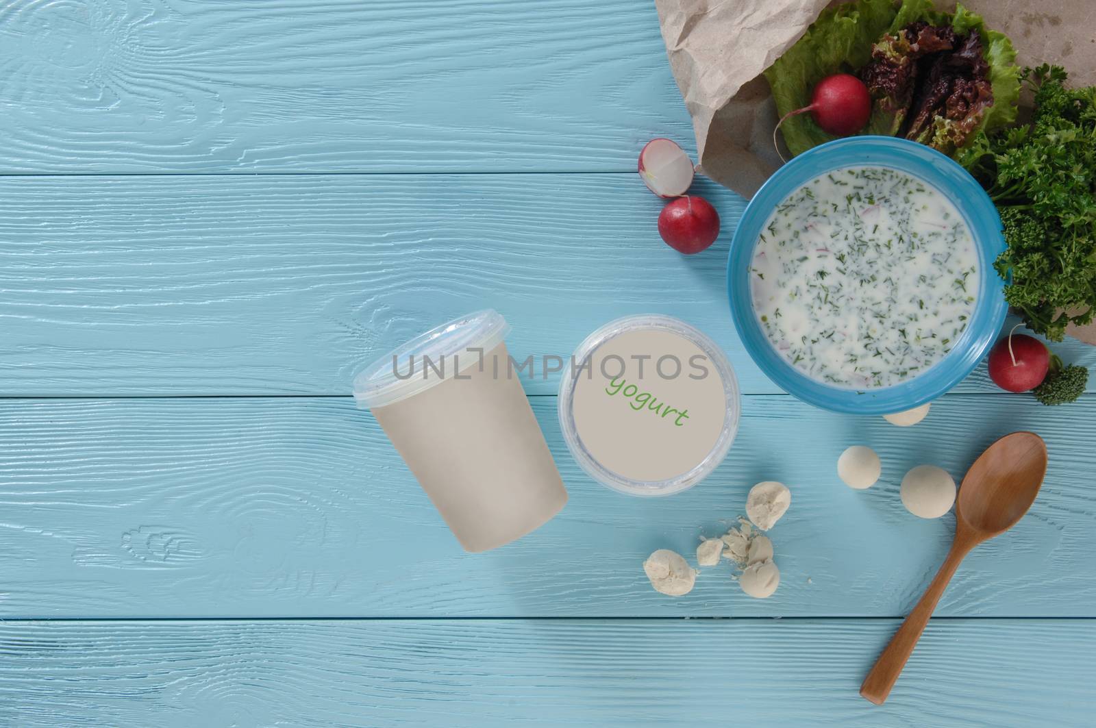 yogurt in a plastic container and greens with radishes on a blue wooden background, top view. healthy eating concept