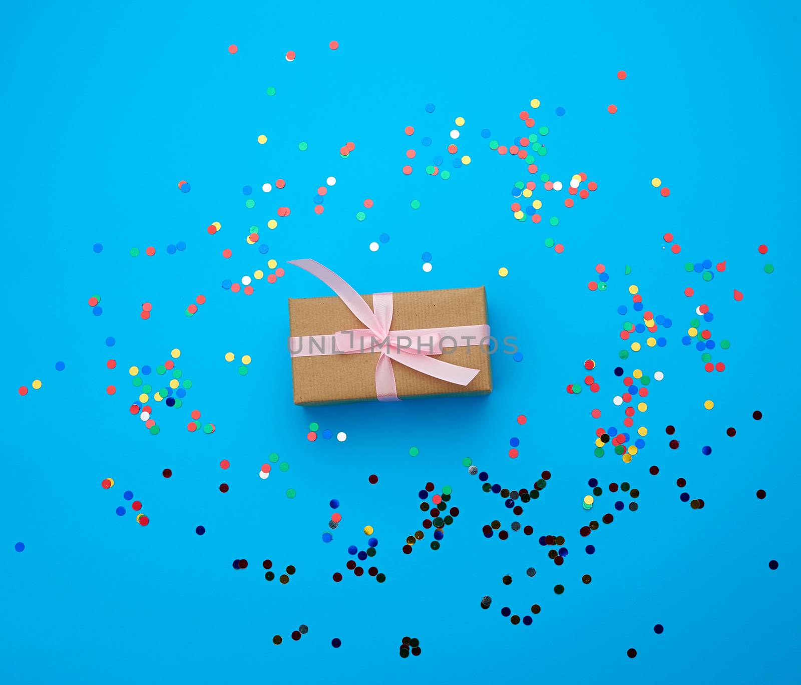 brown gift box wrapped in paper and tied with silk pink ribbon on a blue background with multi-colored shiny confetti, festive backdrop for birthday, valentine's day