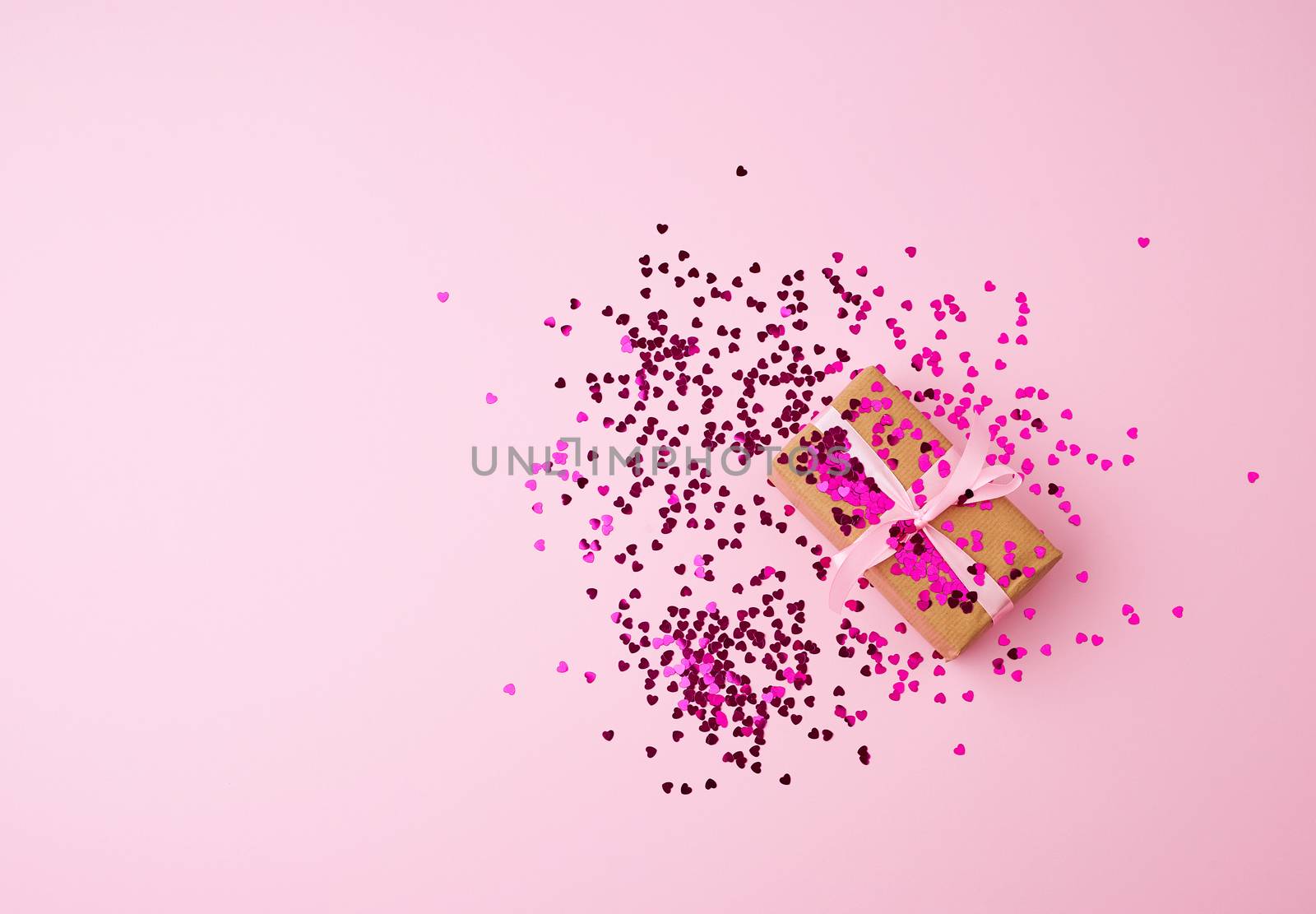 brown gift box wrapped in paper and tied with silk pink ribbon on a pink background with multi-colored shiny confetti, festive backdrop for birthday, valentine's day