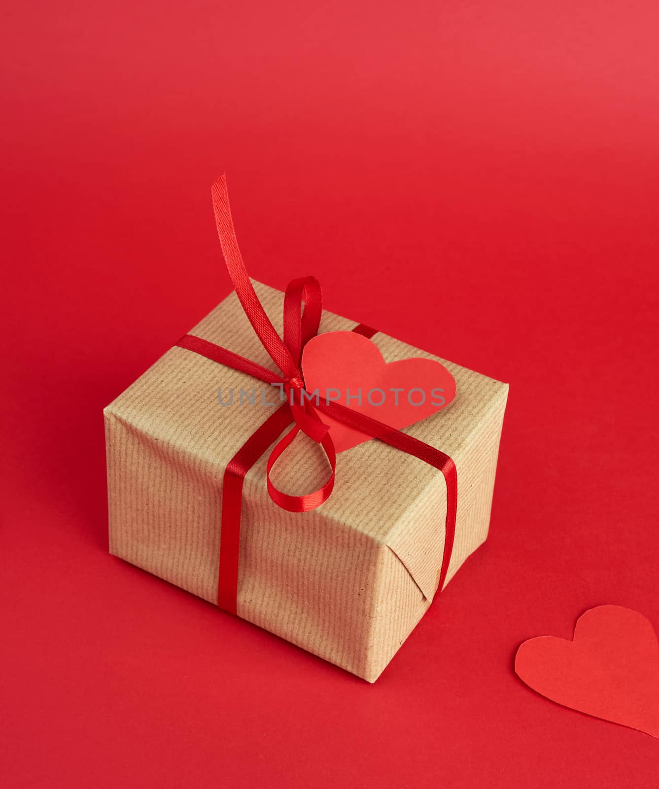 wrapped square box with a gift in brown craft paper and tied with a thin silk red ribbon on a red background, empty paper heart on a gift