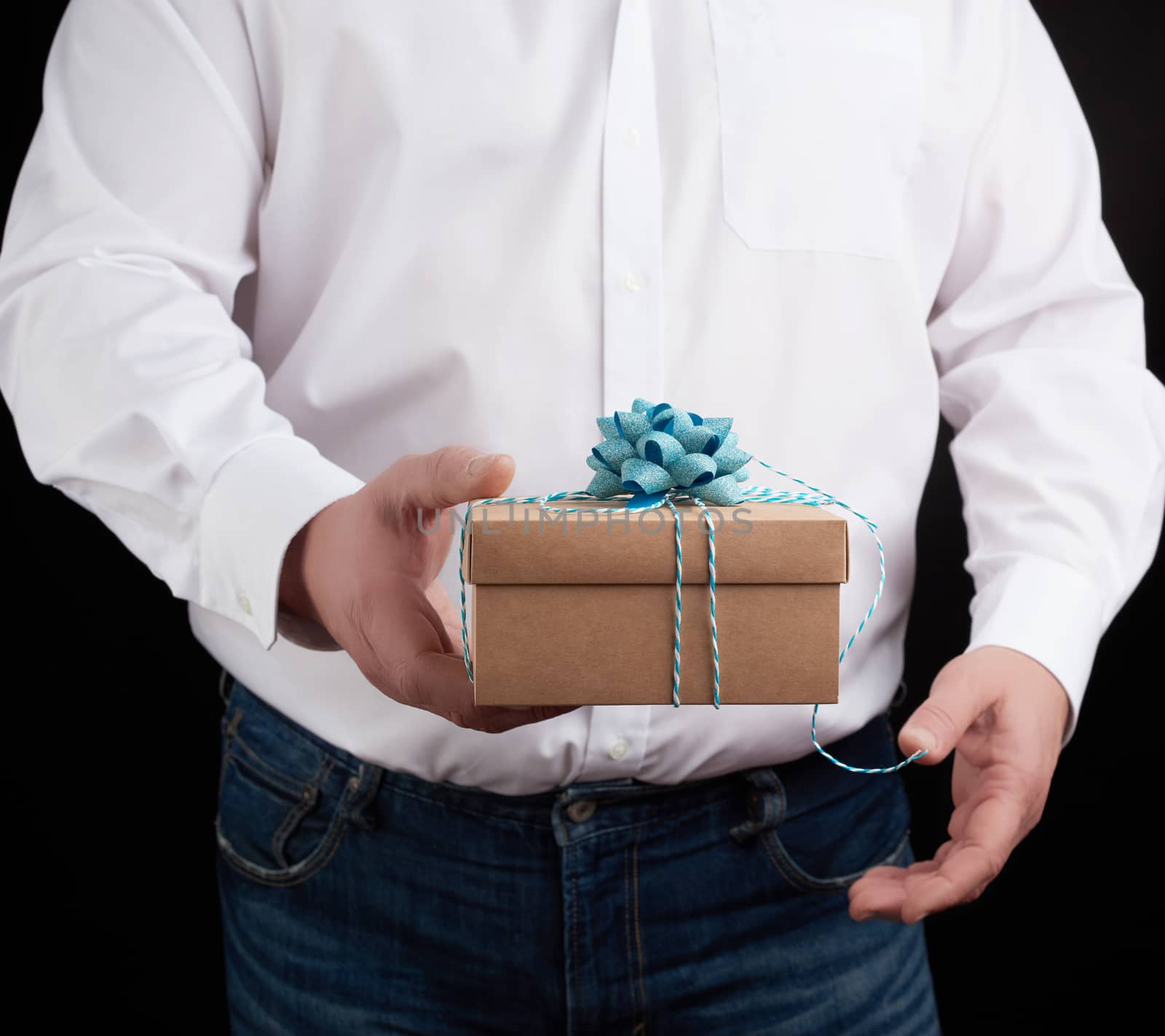 man in a white shirt holds in his hand a closed brown gift box on a dark background, concept proposal of marriage, congratulations on the holiday, copy space