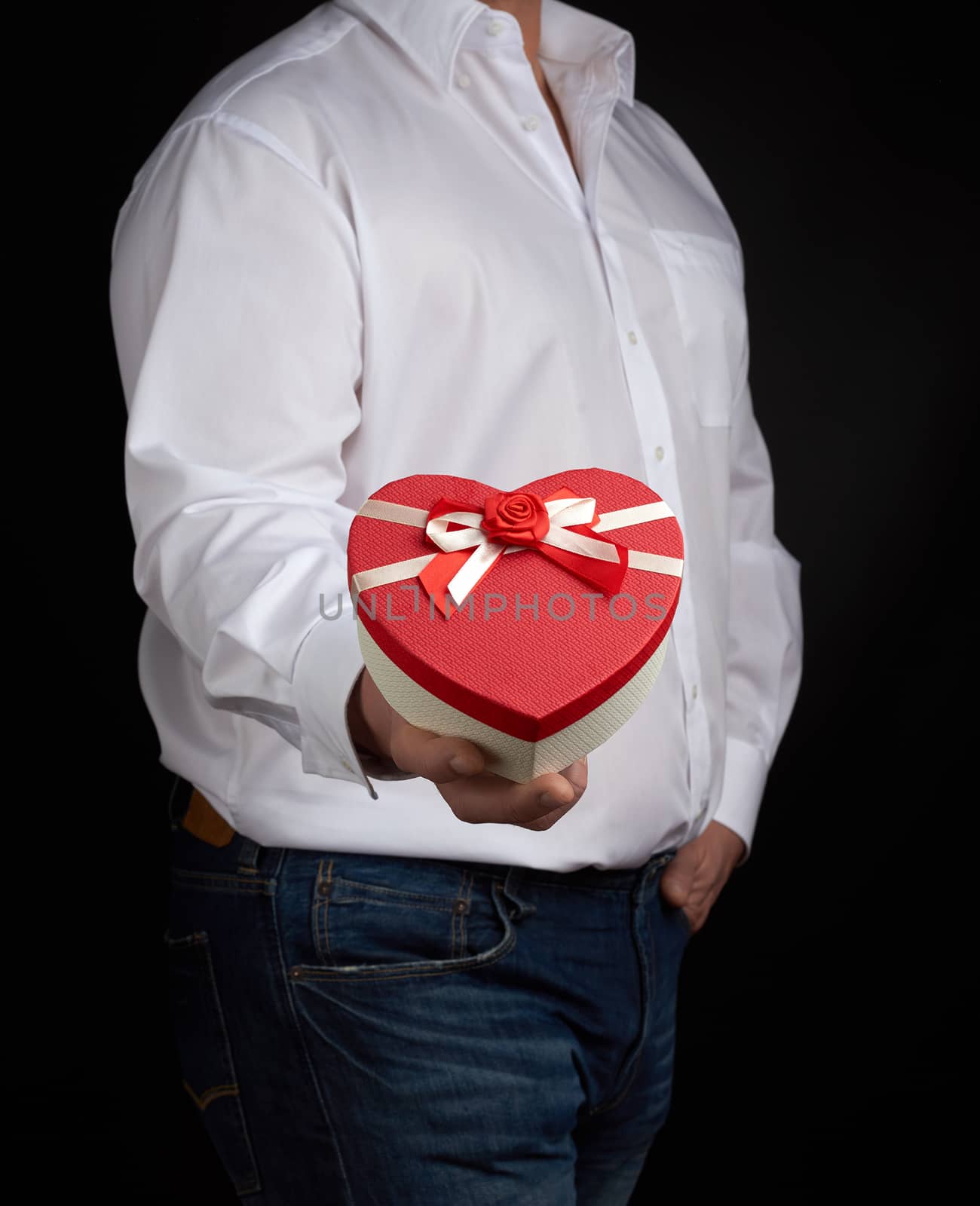 adult man in a white shirt holds a red cardboard box in the form of a heart with a bow on a dark background, concept of holiday, surprise and a gift