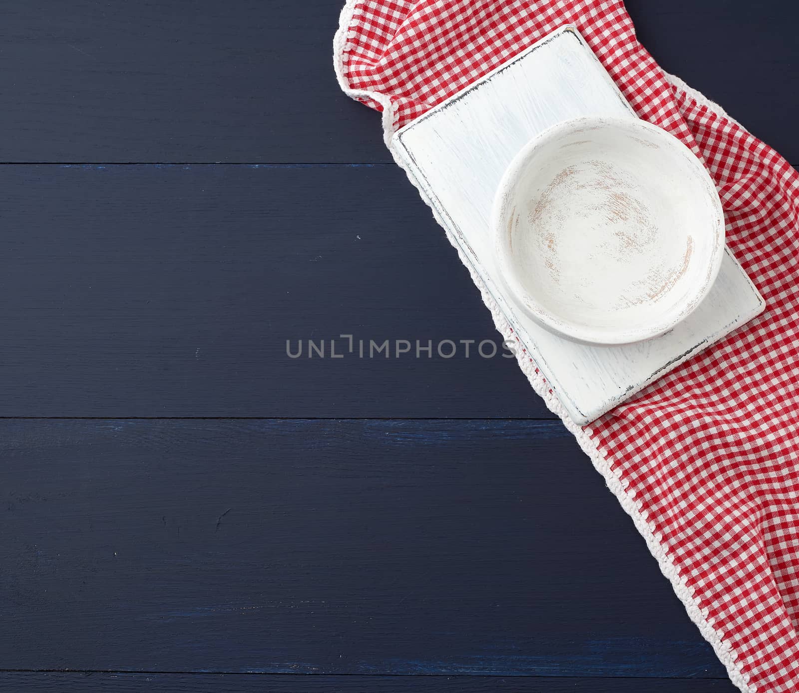 rectangular white cutting kitchen board and empty round plate on a blue wooden background, next to a white red textile towel, top view. Copy space