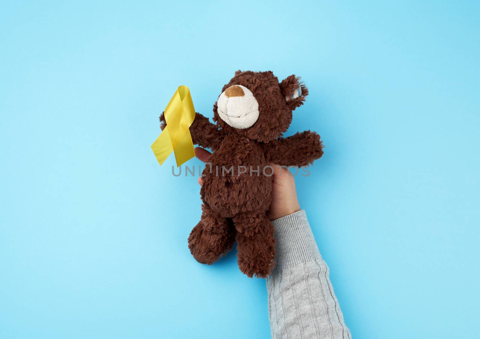 female hand hold a small brown teddy bear which holds in its paw a yellow ribbon folded in a loop on a blue background. concept of the fight against childhood cancer. problem of suicides