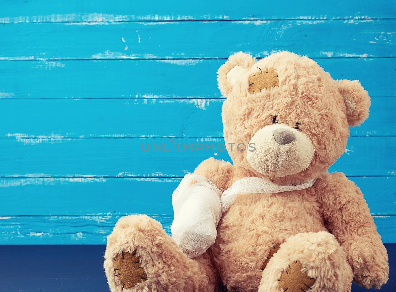 brown teddy bear with rewound white bandage paw on a blue wooden background, pediatrics concept